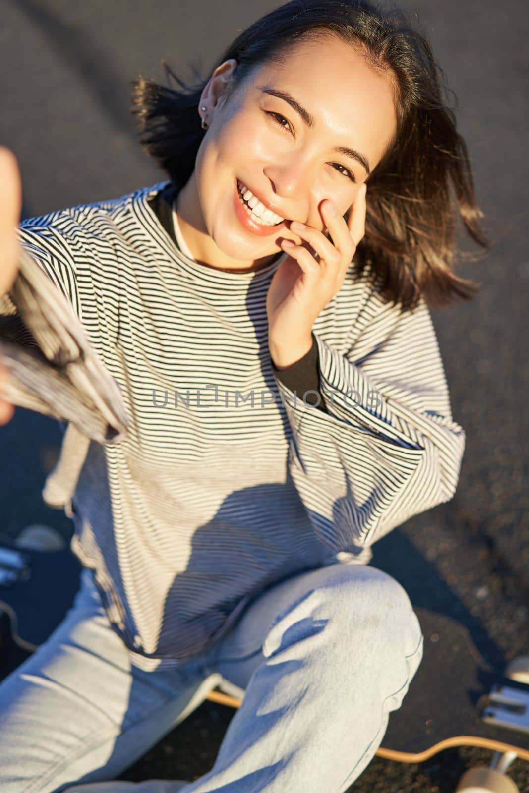 Selfie of beautiful asian girl smiling, taking photo on smartphone while sitting on skateboard outdoors by Benzoix