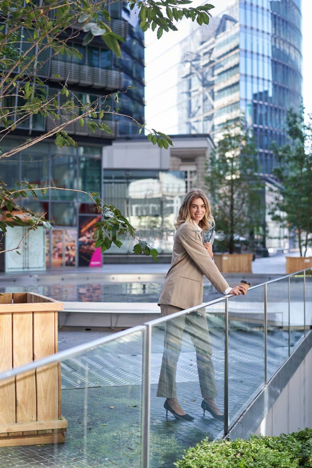 Vertical shot of corporate woman in beige suit standing outside on street, drinks morning coffee takeaway before work by Benzoix