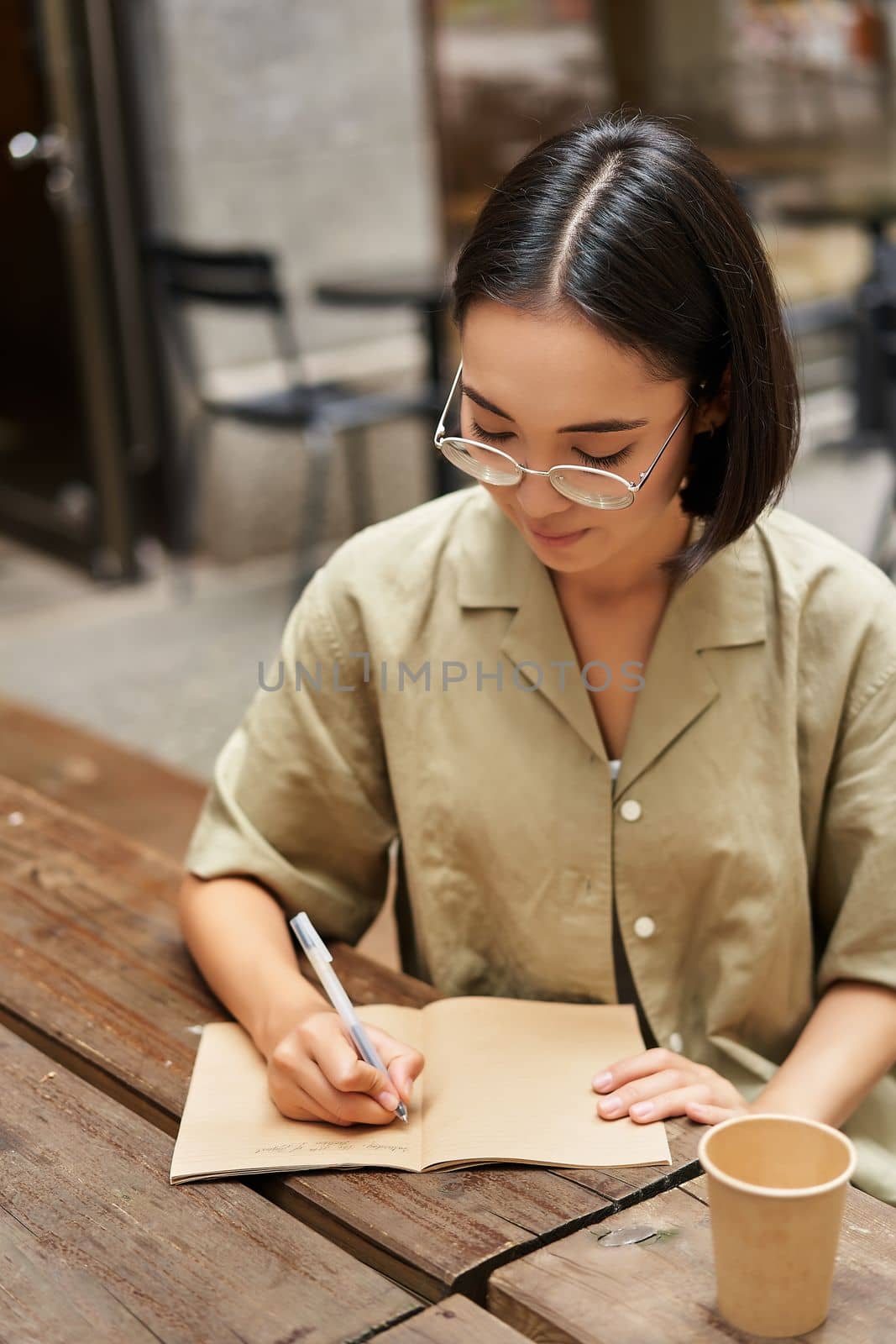 Vertical shot of young asian woman doing homework, making notes, writing something down, sitting in an outdoors cafe and drinking coffee.