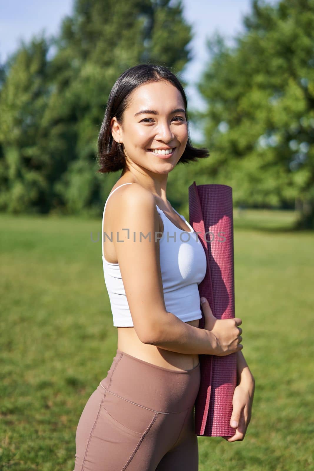 Smiling fitness girl with rubber mat, stands in park wearing uniform for workout and sport activities, does yoga outdoors on lawn.