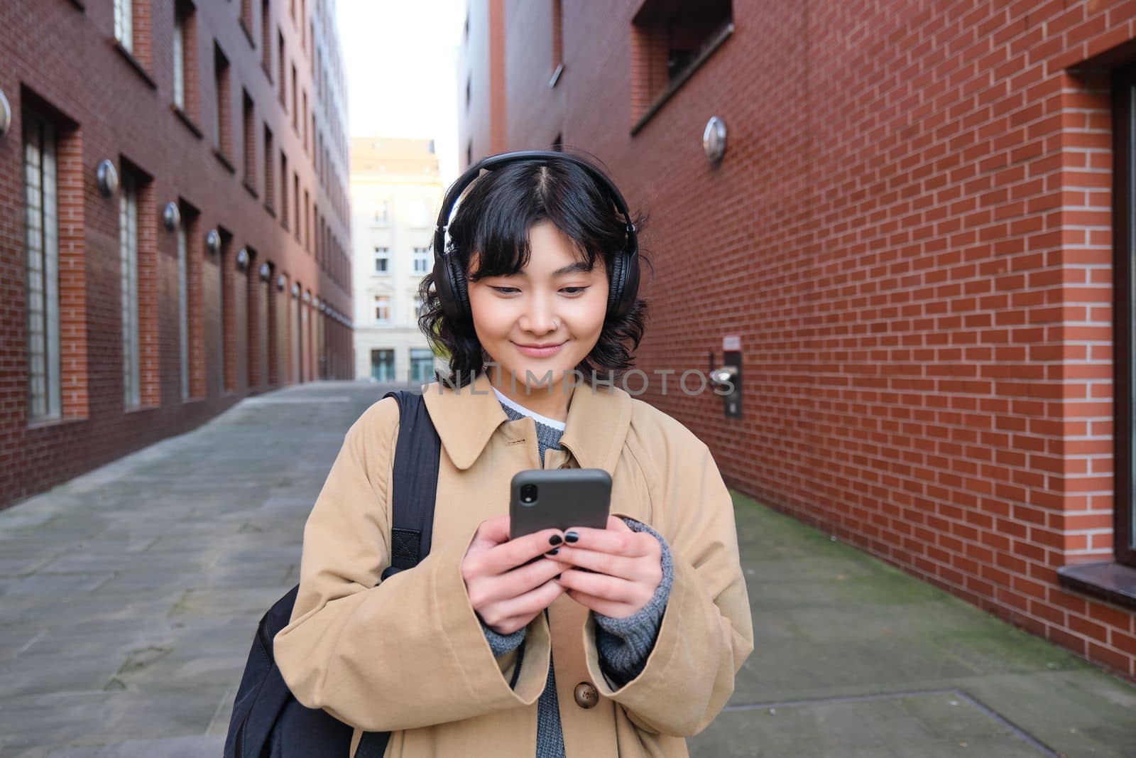 Young asian woman listens music in wireless earphones, uses smartphone on street, walks around city on sunny day, smiling happily.