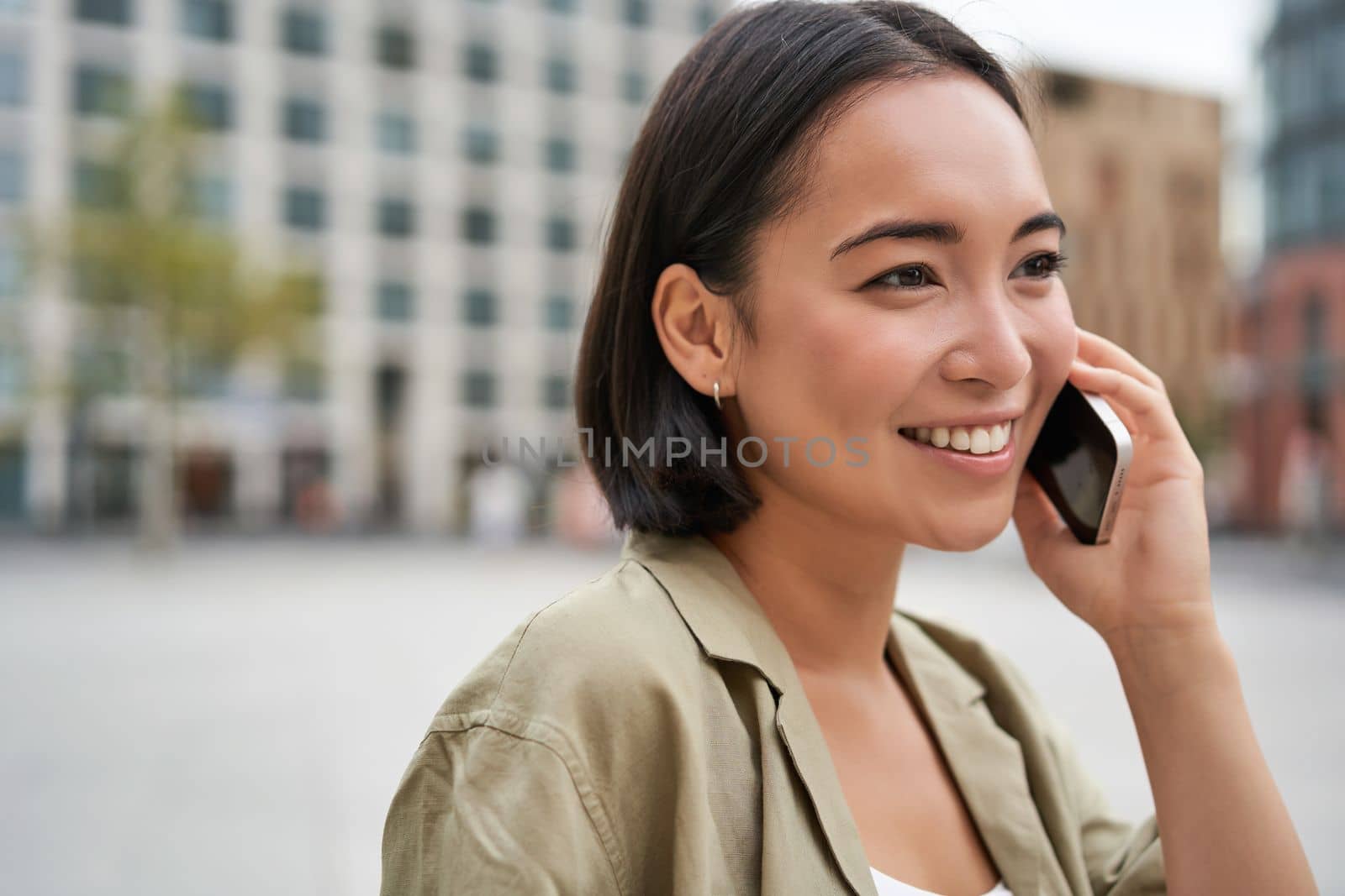 Modern young asian girl talks on mobile phone, uses telephone on city street. Woman smiling while calling someone on smartphone.