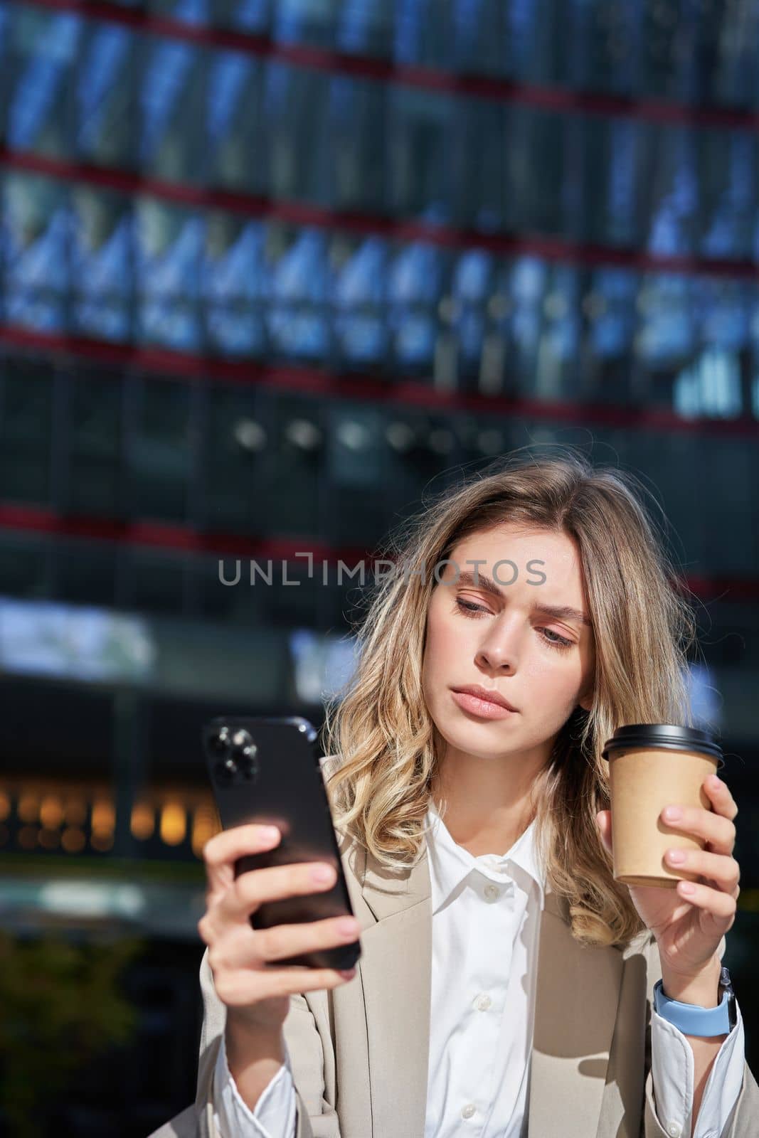 Vertical shot of young corporate woman drinks her coffee and looks at mobile phone, texts message, stands outside in city.
