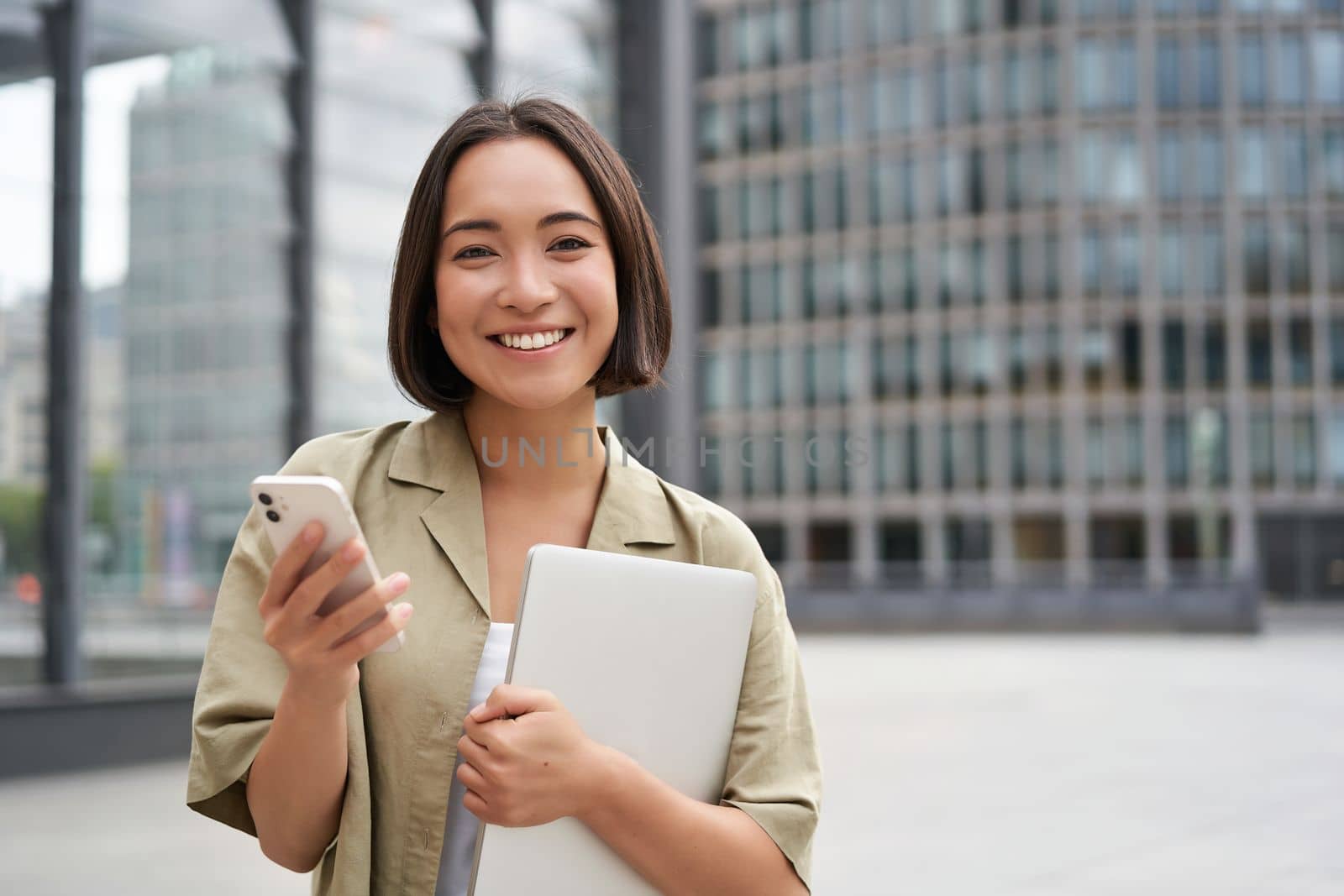 Portrait of smiling asian girl with laptop, holds mobile phone and looks happy at camera, stands on street.