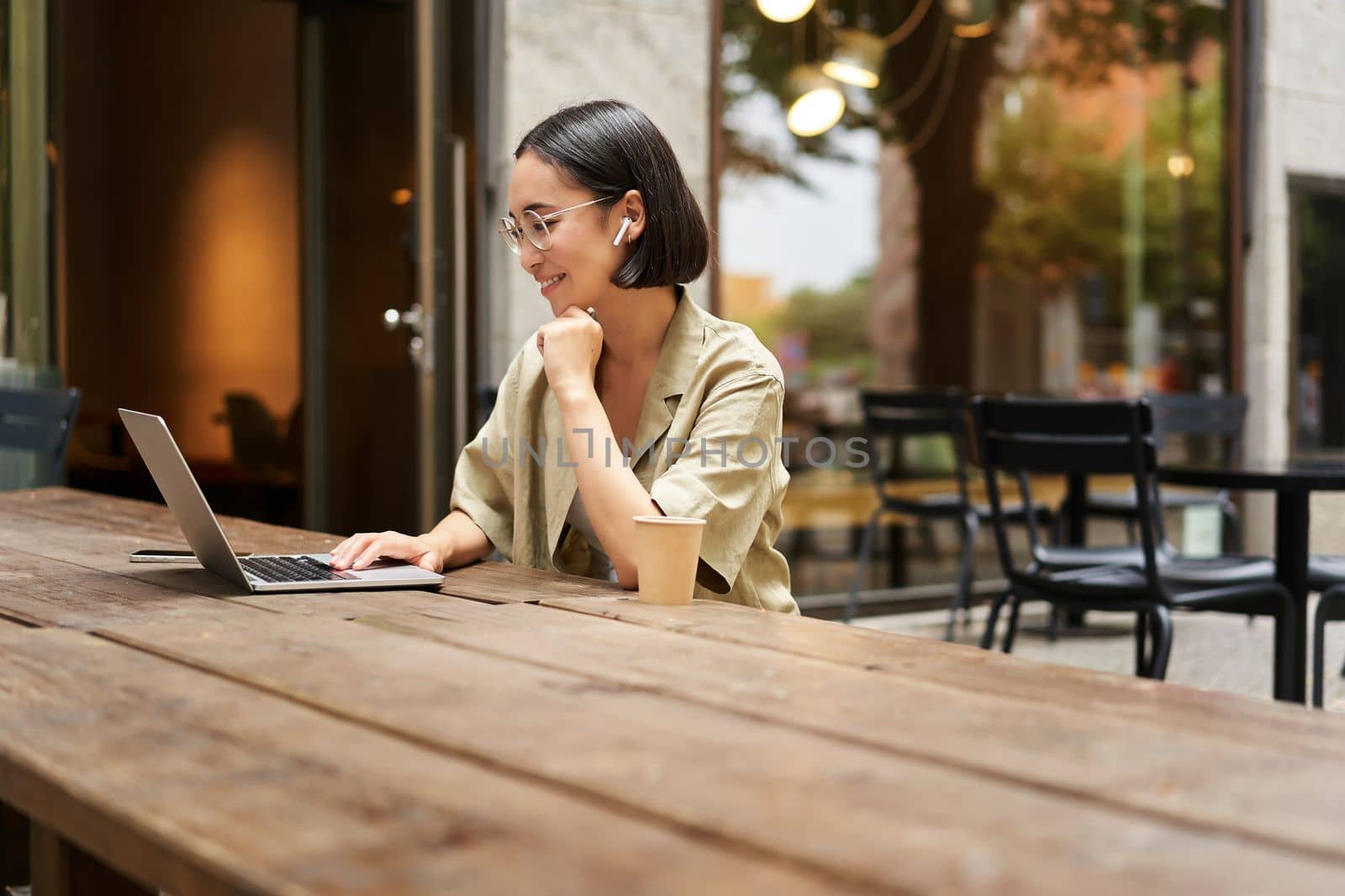 Silhouette of young woman working in cafe with laptop, typing on keyboard and drinking coffee, sitting outside.