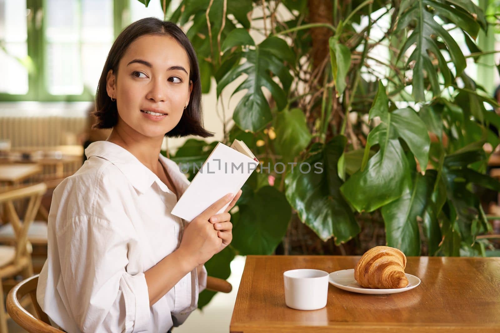 Beautiful young asian woman with a book in hands, sitting in cafe, drinking coffee and eating croissant, smiling, looking mysterious.