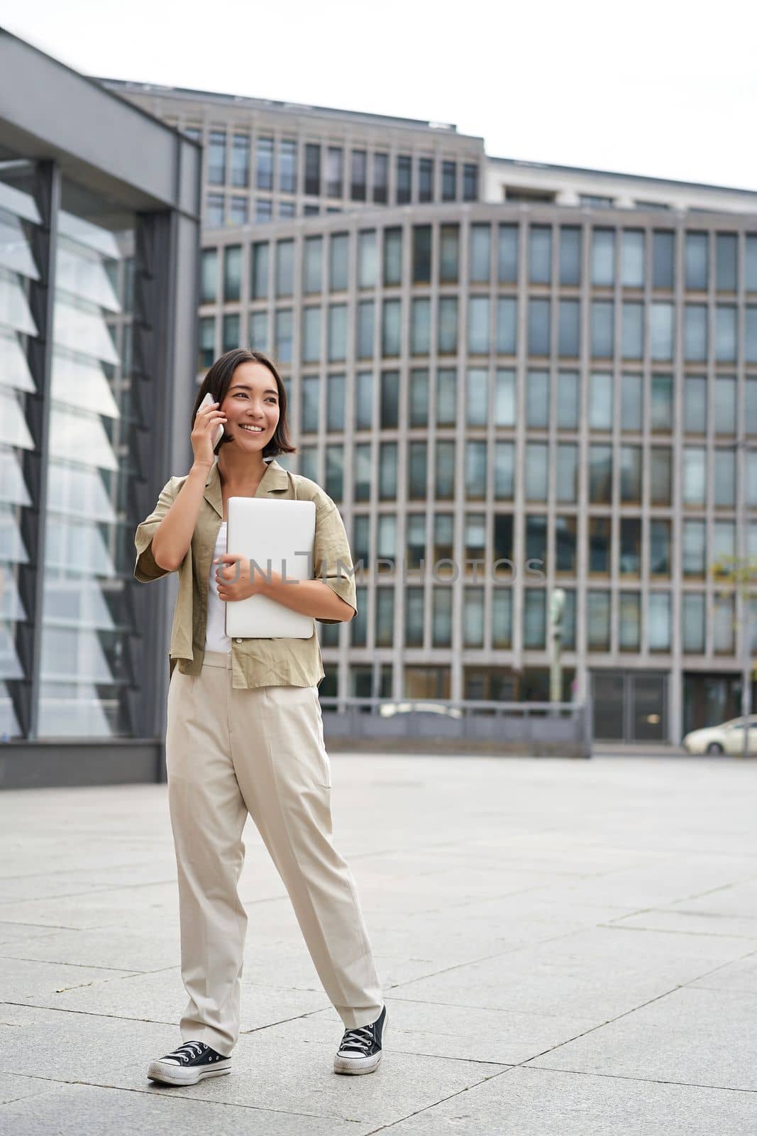 Stylish urban girl walking on street, talking on mobile phone and carrying laptop. Young woman making a telephone call while going somewhere by Benzoix