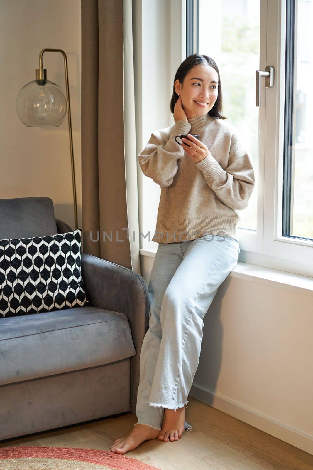 Vertical shot of beautiful woman sitting in her house, looking outside window and drinking coffee.