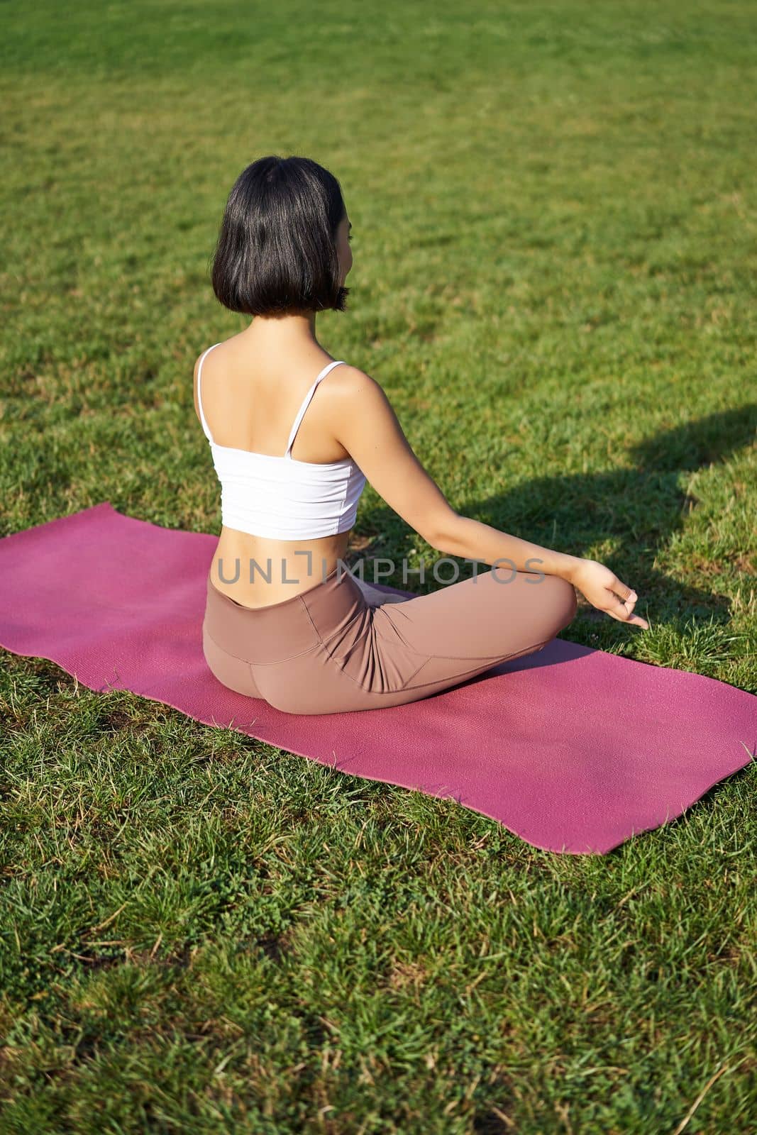 Rear view of woman silhouette doing yoga, sitting on fitness mat and meditating on green lawn.