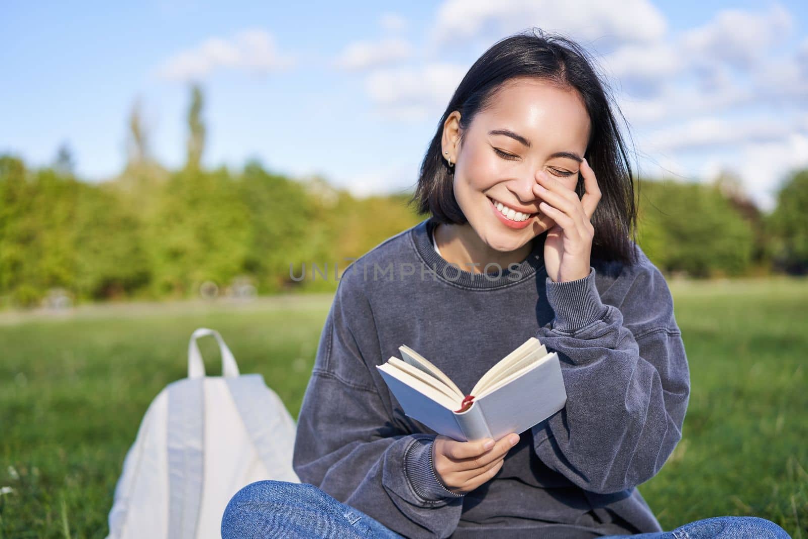 Beautiful woman sitting in park with book. Smiling asian girl reading and laughing, relaxing outdoors.
