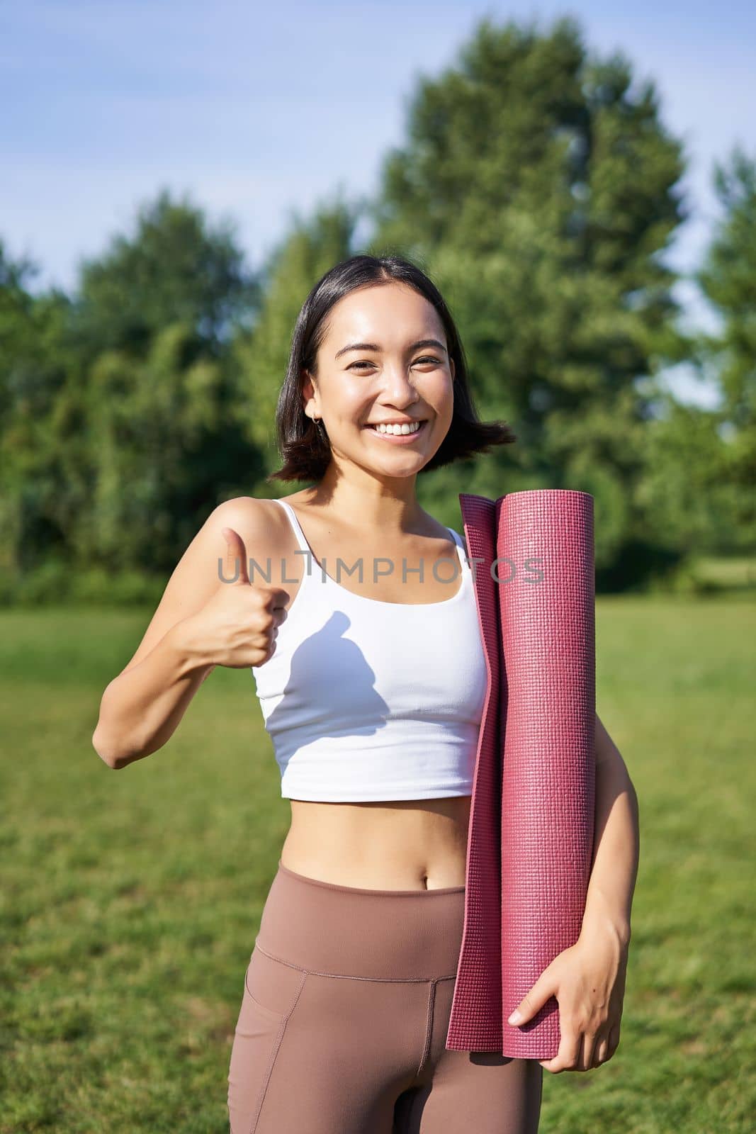 Beautiful and fit fitness girl in leggings, shows thumb up in park, stands with yoga rubber mat for workout and sport training by Benzoix