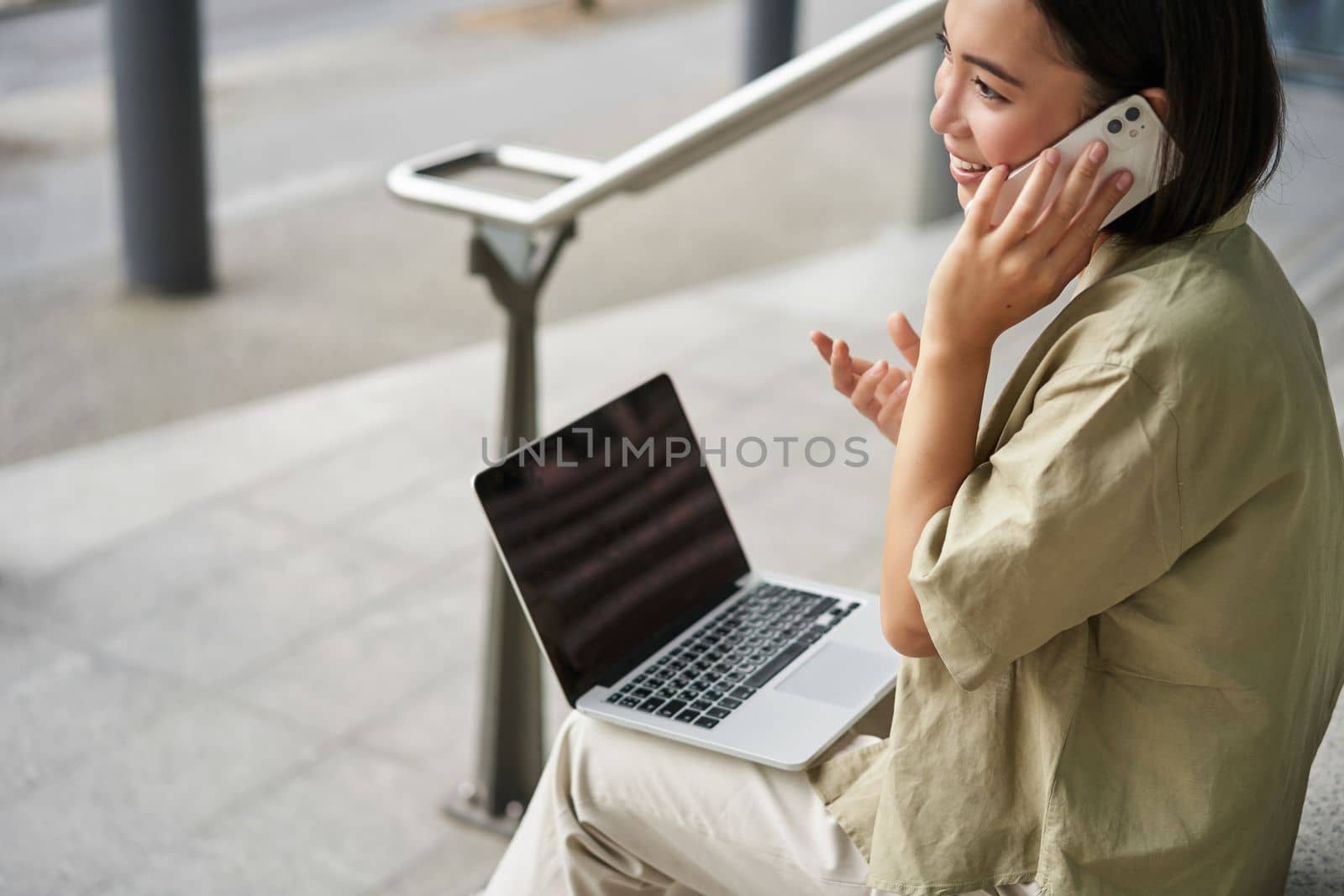 Smiling asian woman makes a phone call. Girl student using laptop and mobile phone, talking to someone on telephone.