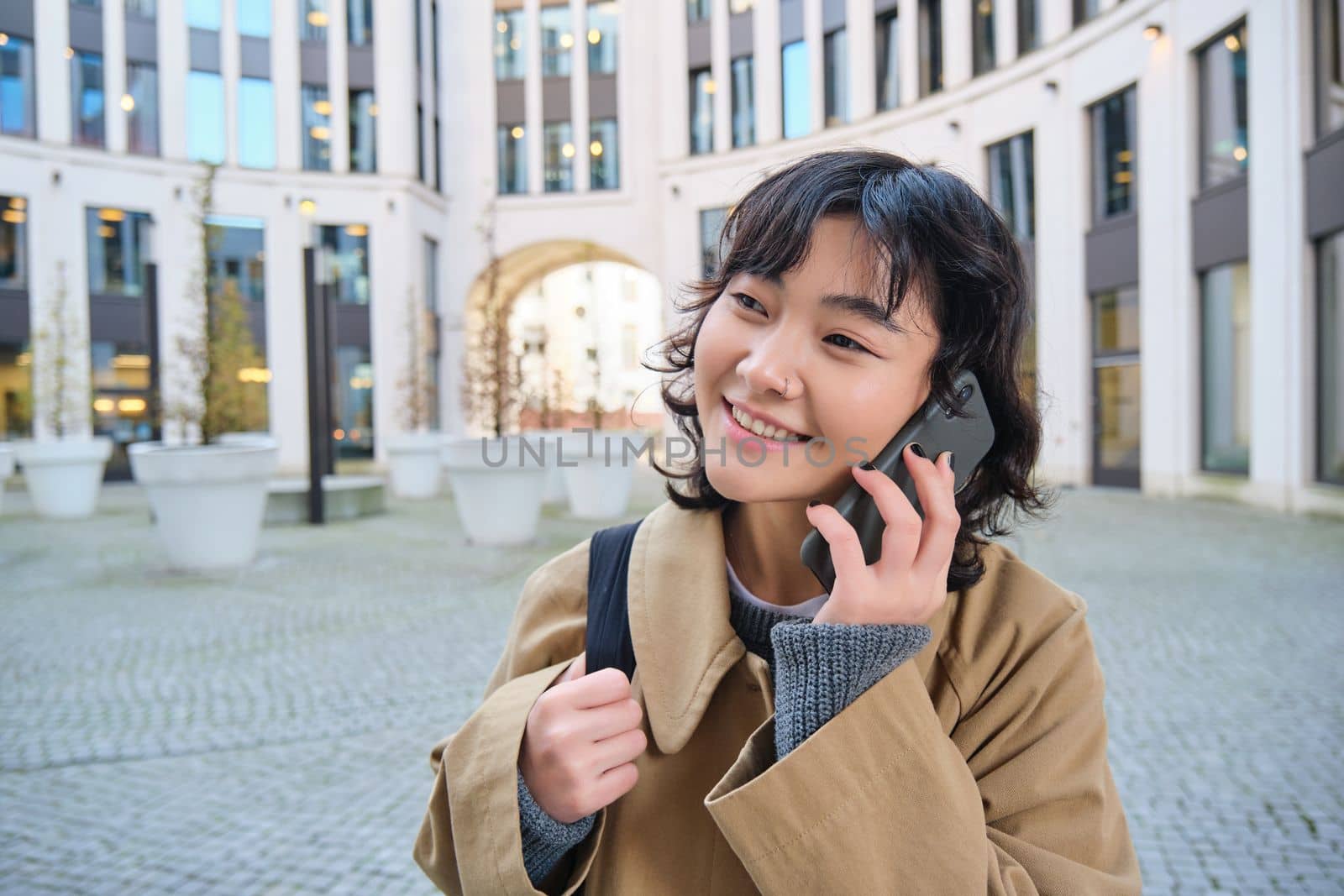 Cellular technology. Young korean woman talks on mobile phone, makes a phone call on her way home, walks down street, city centre, has telephone conversation.