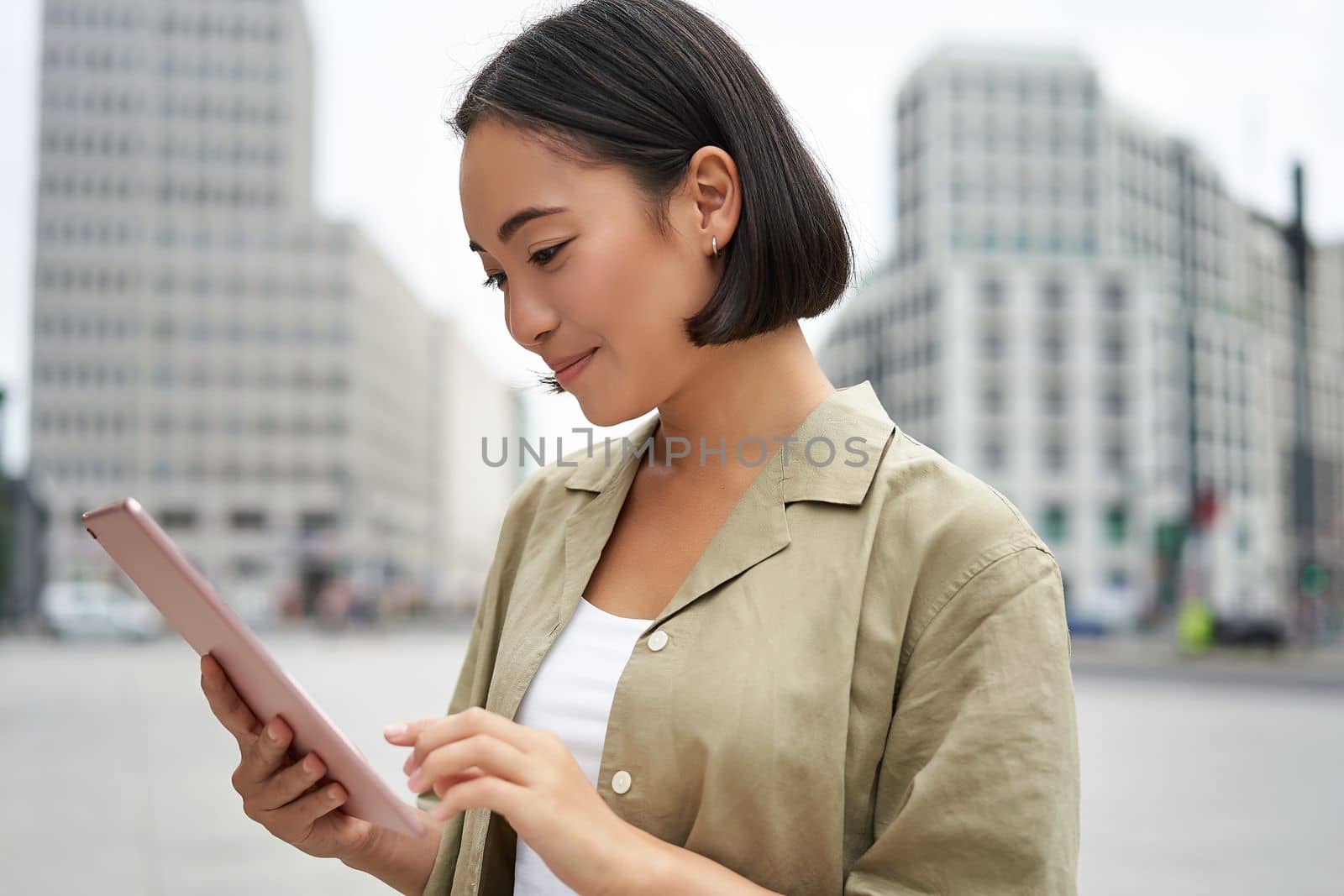 Portrait of asian woman reading, using tablet while standing on street, smiling while looking at screen.
