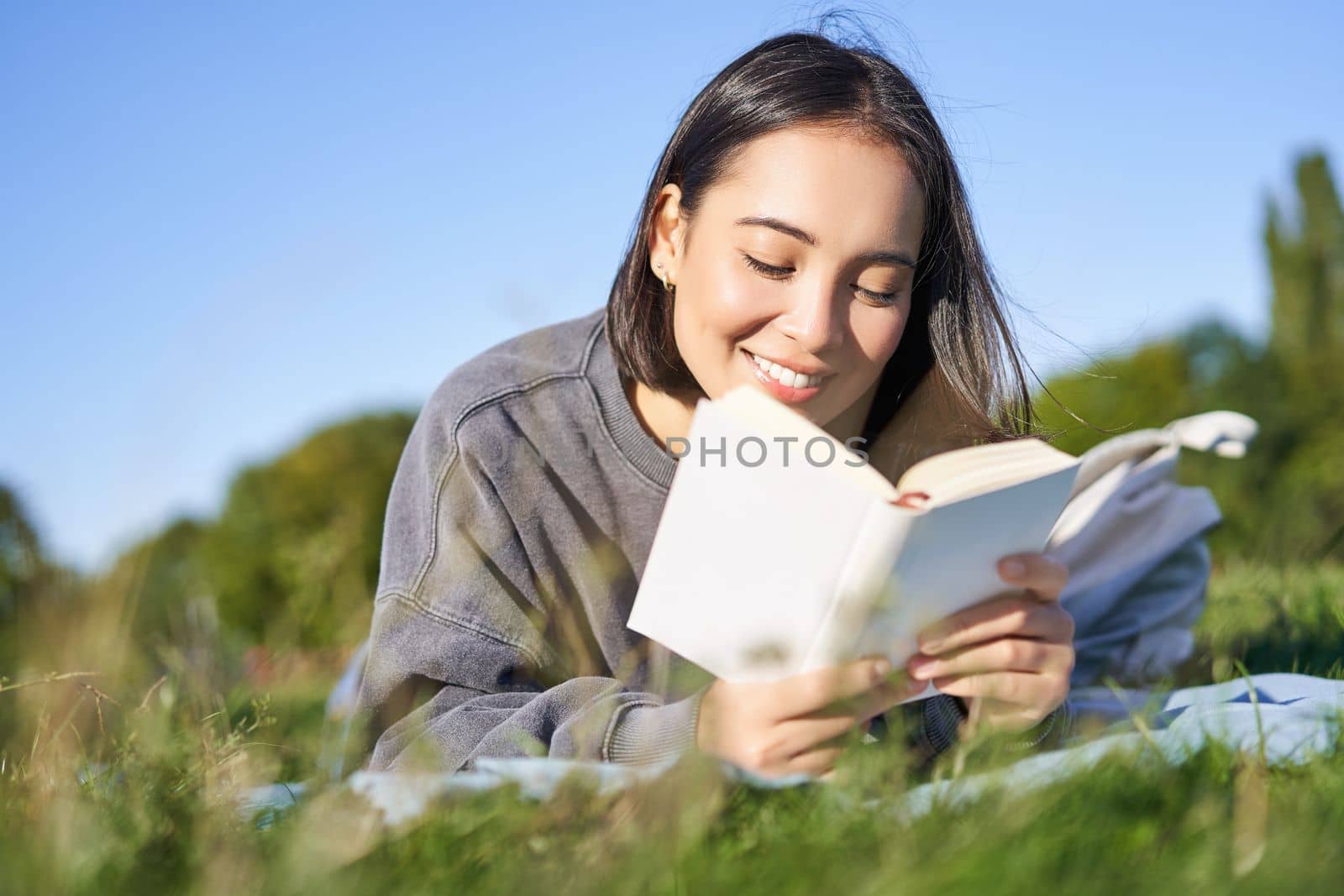Portrait of cute korean girl, reading in park while lying on grass, relaxing with favorite book in hands, smiling happily by Benzoix