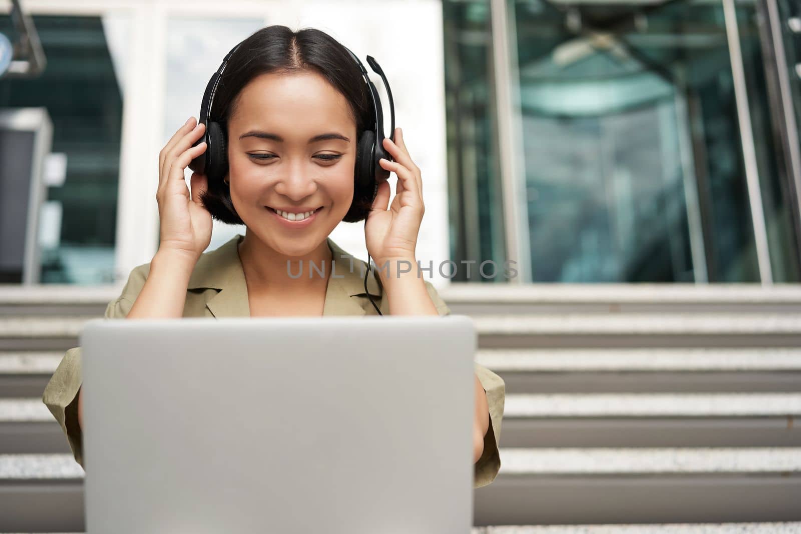 Portrait of young asian woman sitting with laptop and headphones, watching video, does online course on computer, sitting on stairs outdoors by Benzoix