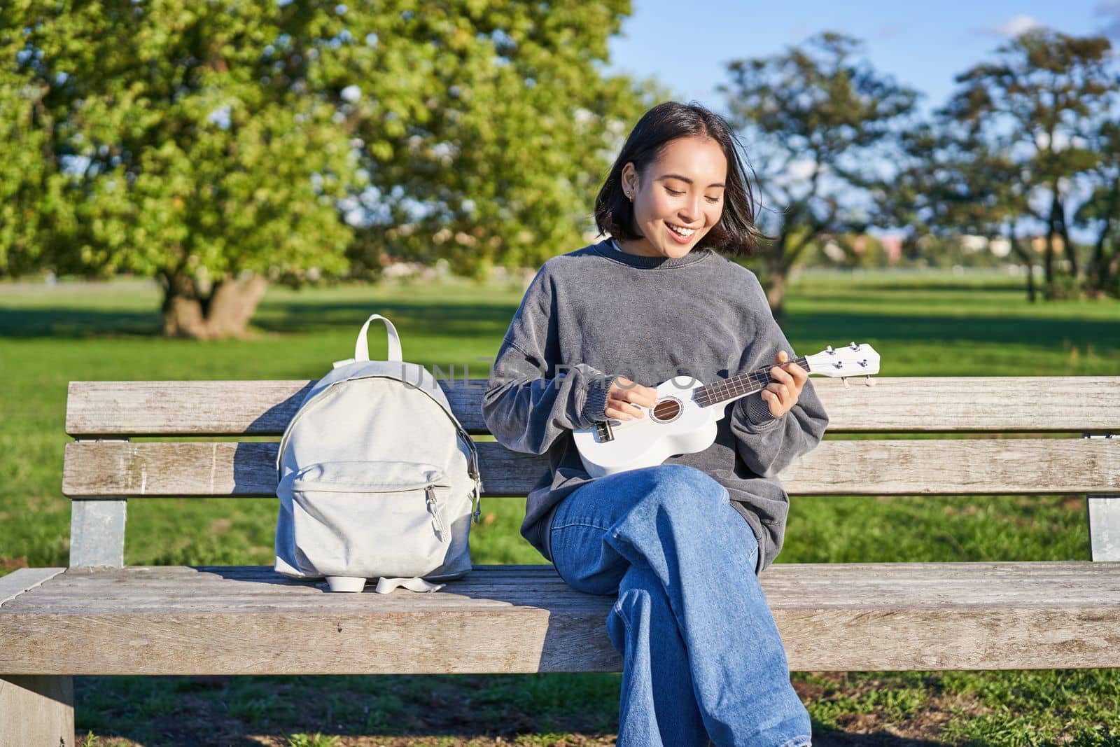 Beautiful asian girl plays ukulele outdoors, sits in park on bench with musical instrument.