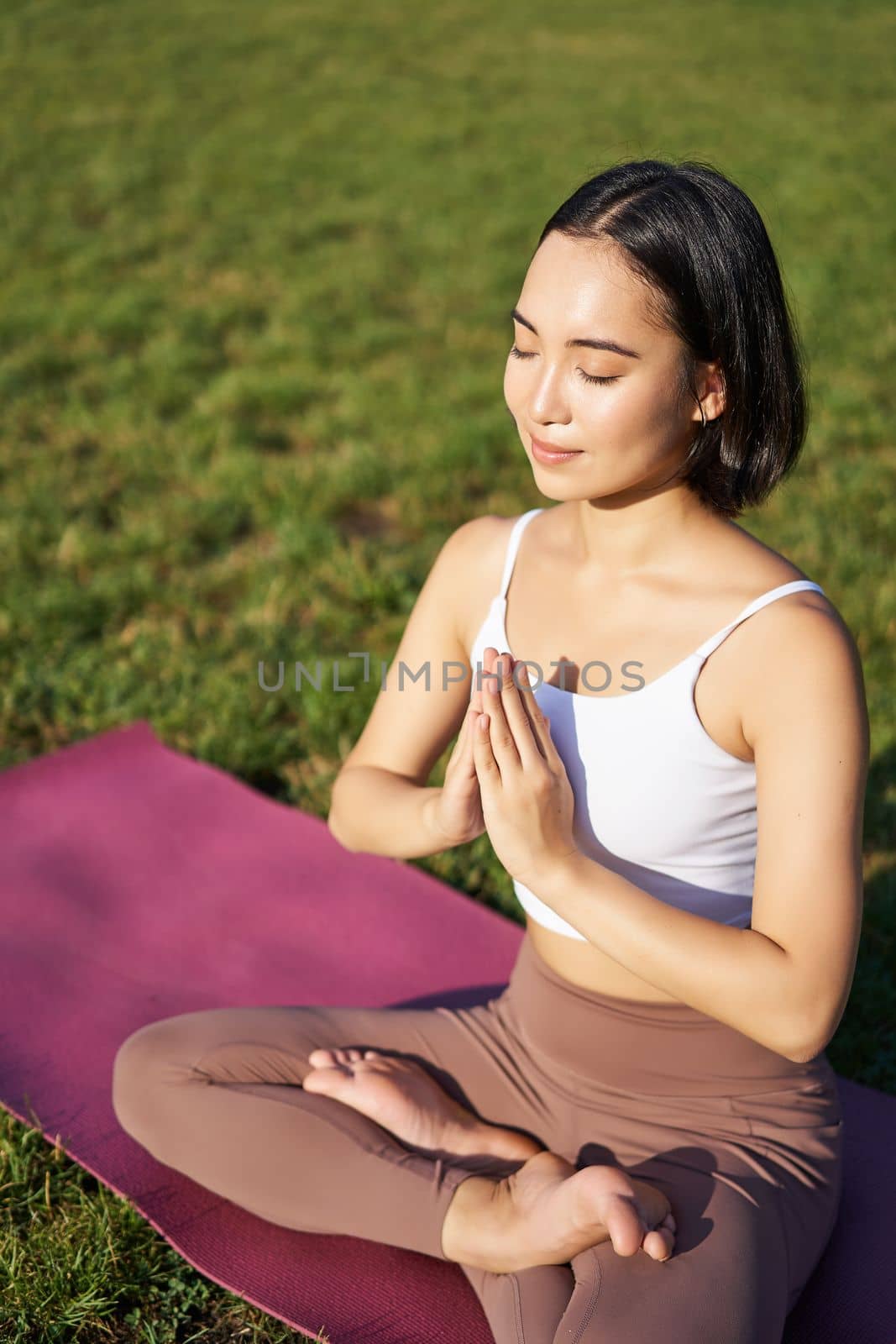 Vertical shot of young asian woman doing yoga, practice mindfulness, smiling and looking relaxed, sitting on rubber mat in park by Benzoix