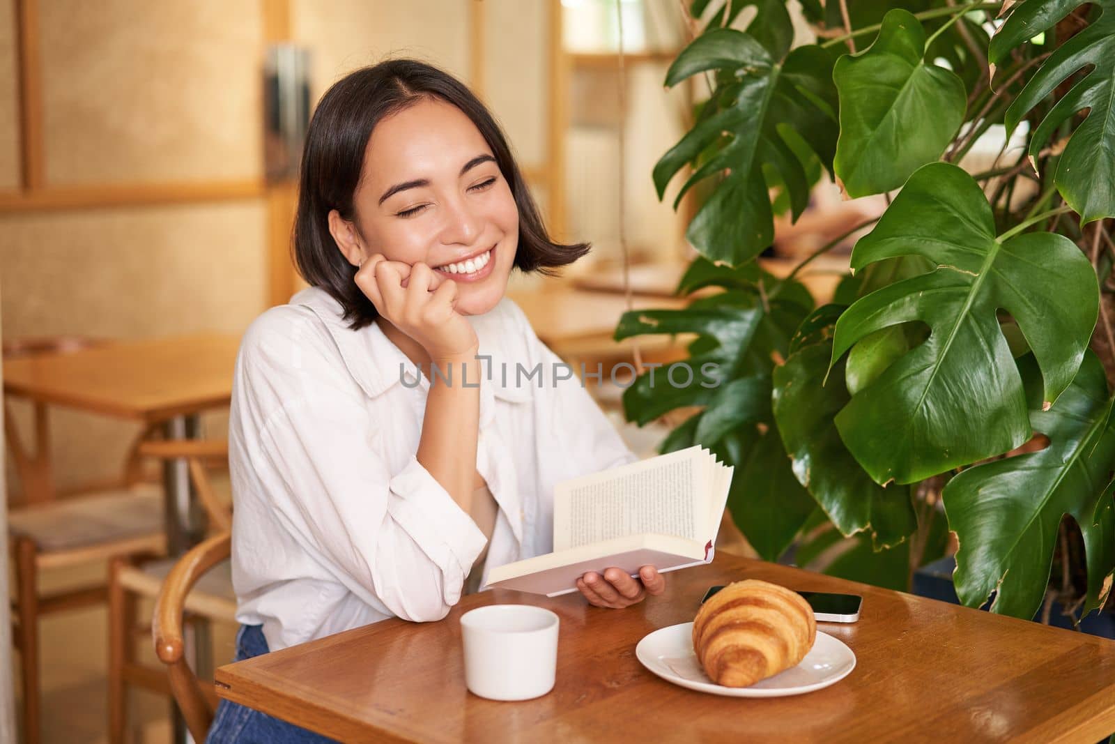 Romantic asian woman sitting with book in cafe, eating croissant and drinking coffee, reading and smiling, enjoying alone time.