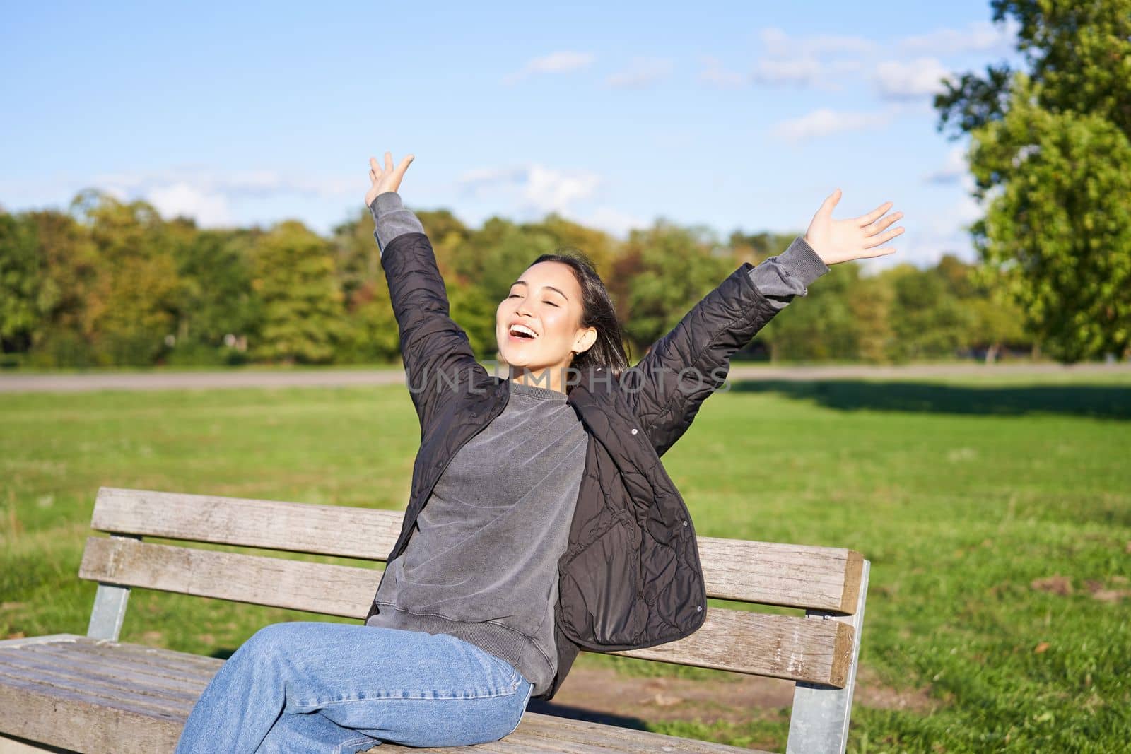 Happy asian woman stretching her hands, sitting on bench with excited face, smiling pleased, feeling freedom, enjoying day in park by Benzoix
