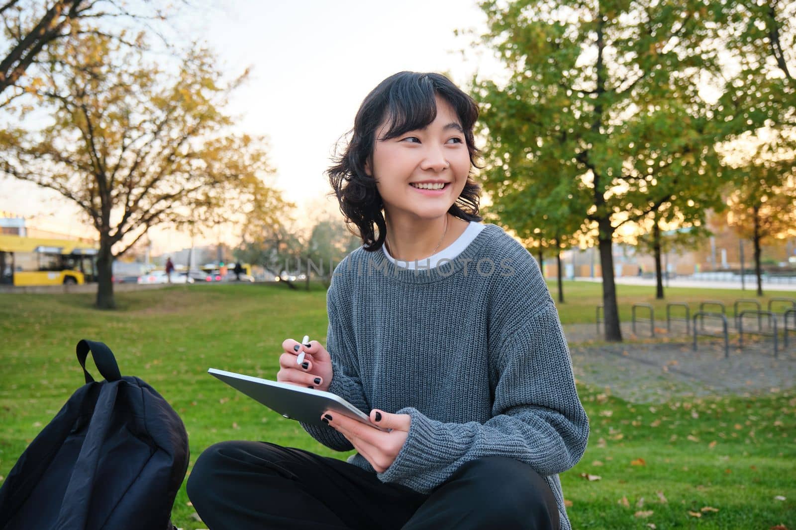 Young korean woman sits in park, draws on digital tablet, writes in gadget with pen tool, looks around and makes notes, creates artwork or scatches.