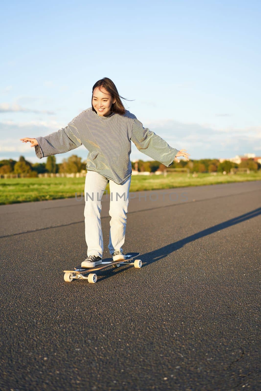 Cute asian girl riding skateboard, skating on road and smiling. Skater on cruiser longboard enjoying outdoors on sunny day.