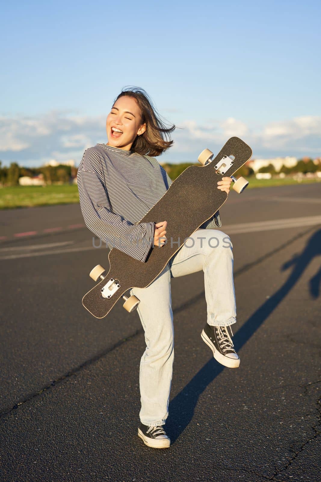 Funny asian girl enjoying skating, holding skateboard like guitar and shadow playing, having fun outdoors.