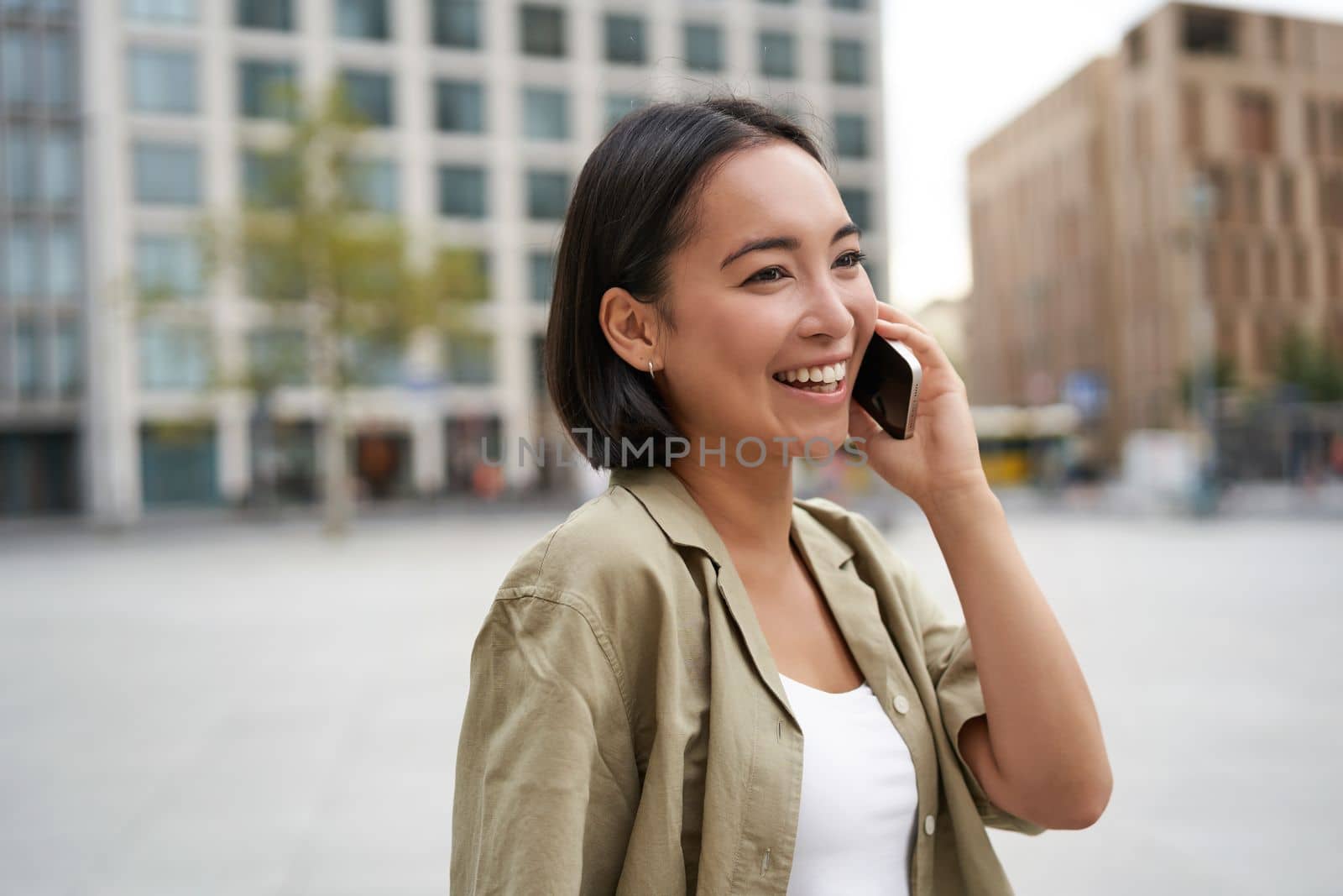 Modern young asian girl talks on mobile phone, uses telephone on city street. Woman smiling while calling someone on smartphone.