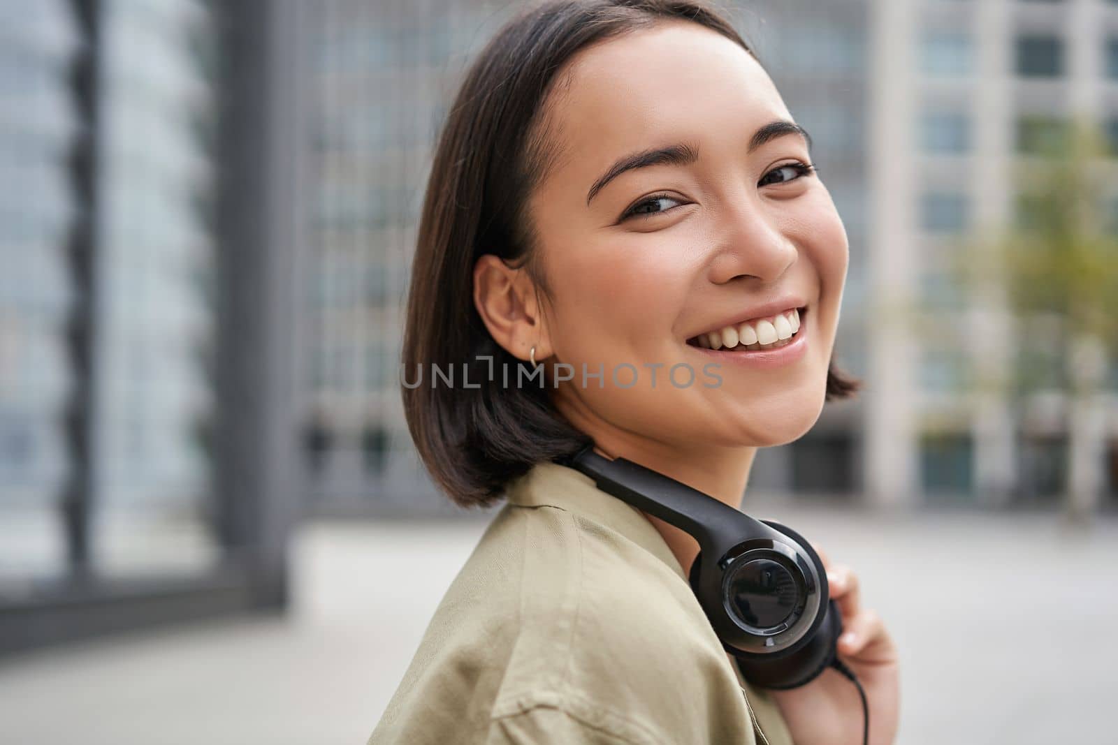 Portrait of happy asian woman in headphones, enjoys music while walking in city, smiling and laughing.