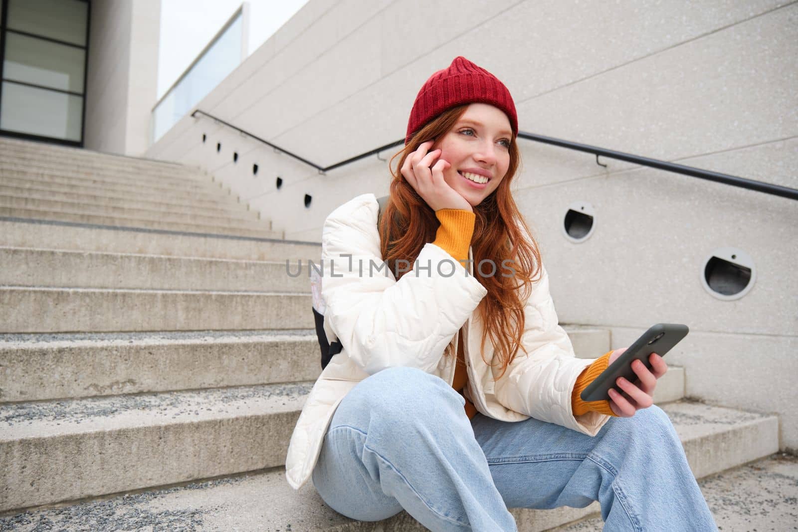 Stylish european girl with red hair, sits on public stairs with smartphone, places online order, sends message on mobile phone social app, smiles happily.