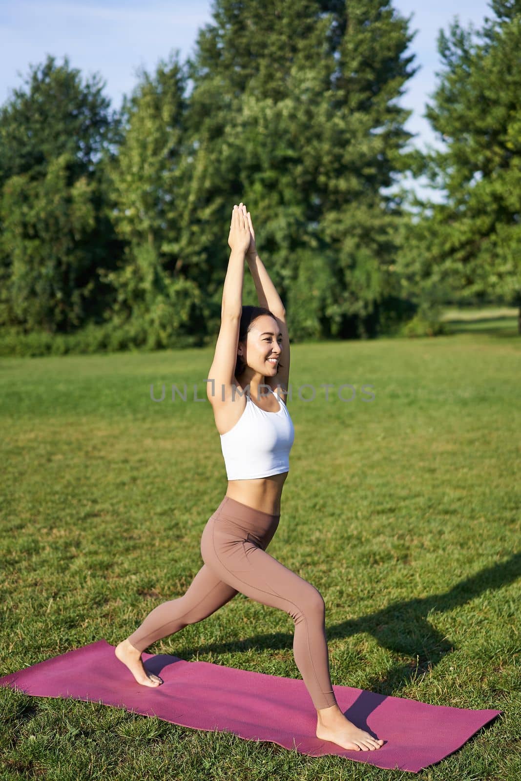 Vertical shot of asian woman standing in tree pose, making yoga asana on fresh air, wellbeing training in park on rubber mat.