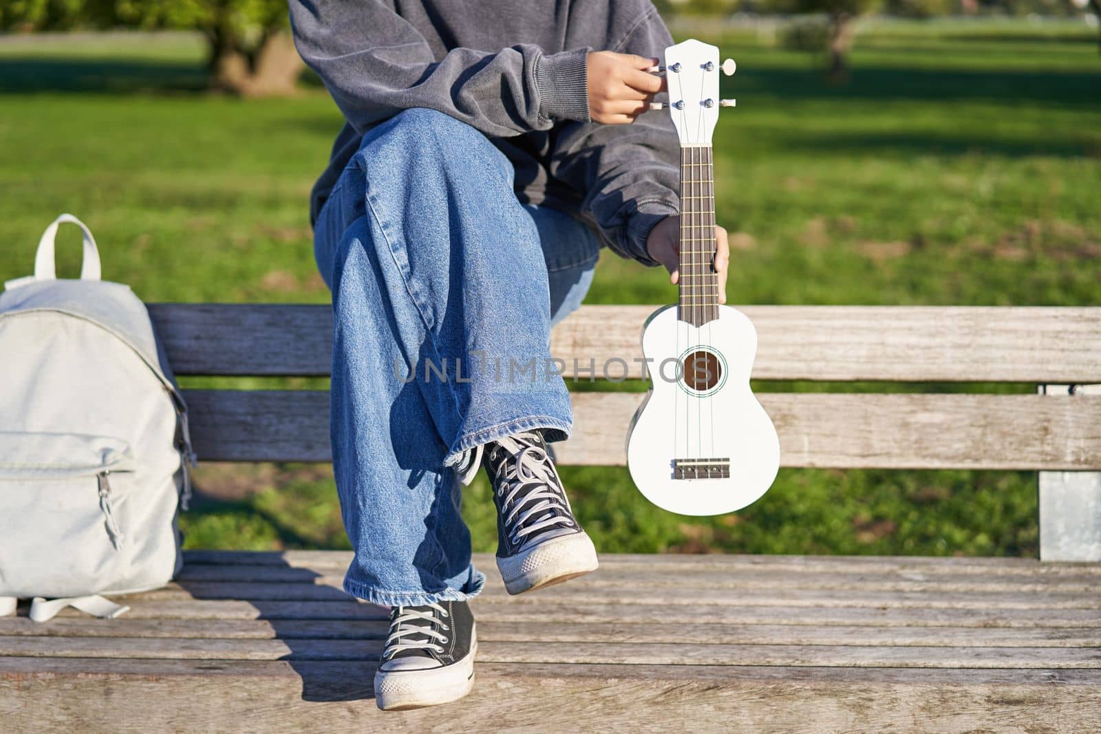 Cropped shot of young girl in sneakers and jeans, hands holding ukulele musical instrument while she sits on bench in green sunny park.