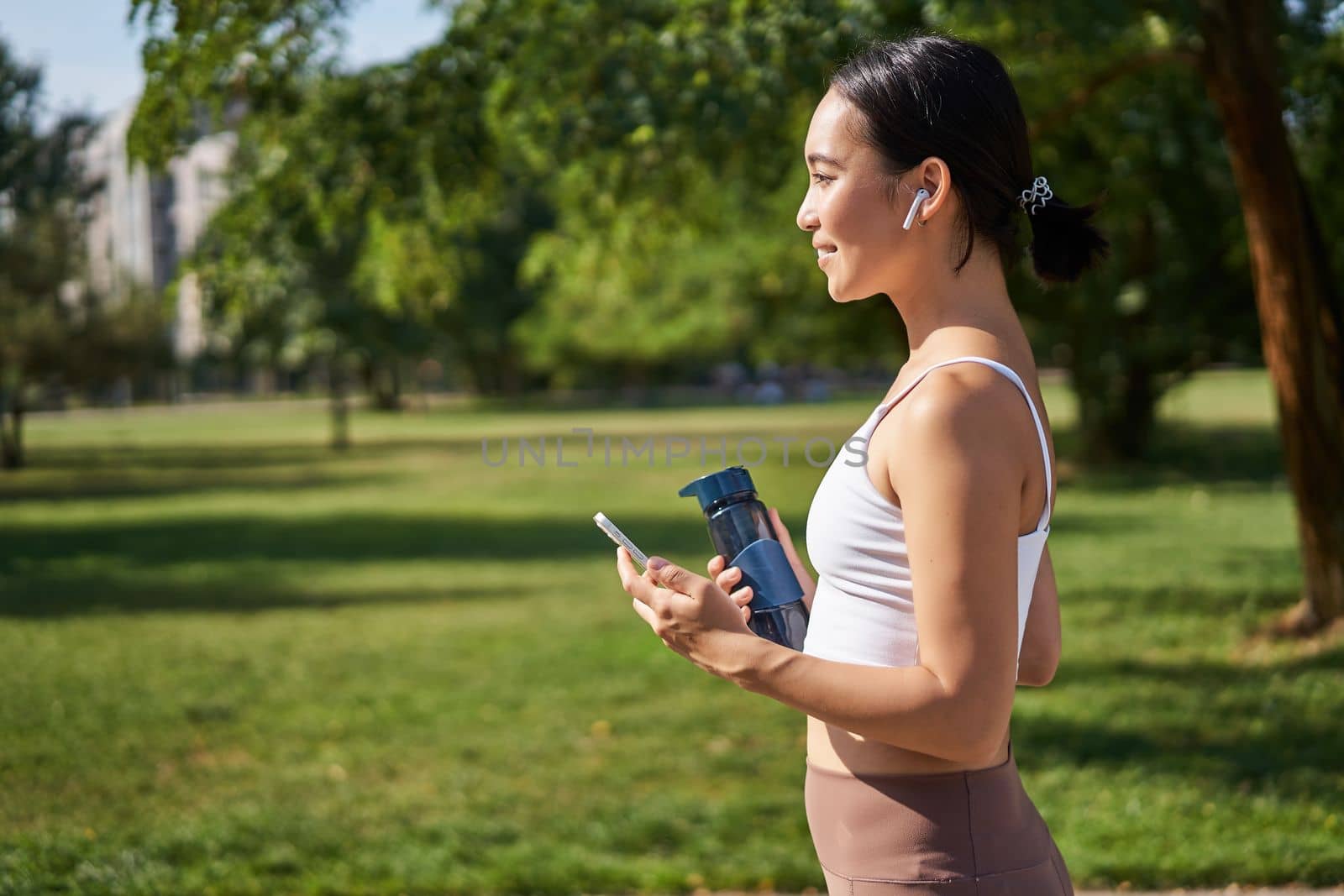 Active asian girl, in fitness clothing, workout in park, walking in sportswear with smartphone and water bottle by Benzoix