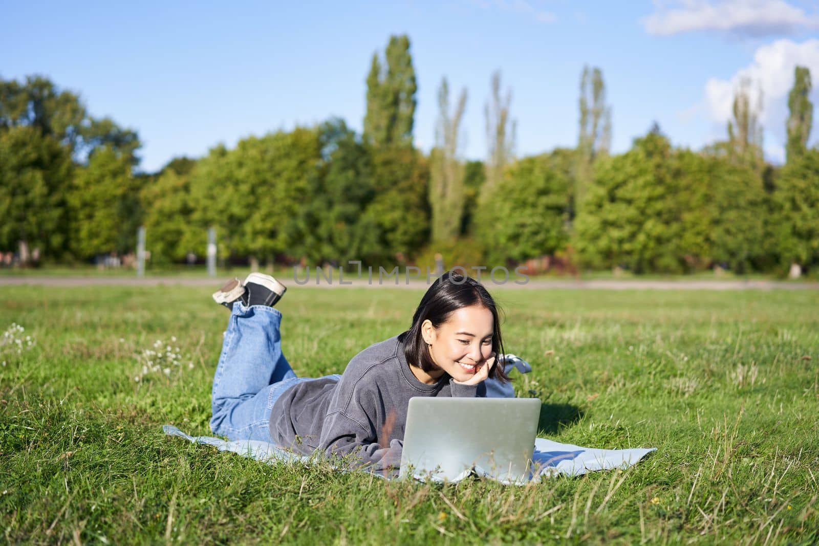 Portrait of young happy asian girl lying on blanket in park, watching videos and browsing internet on her laptop.