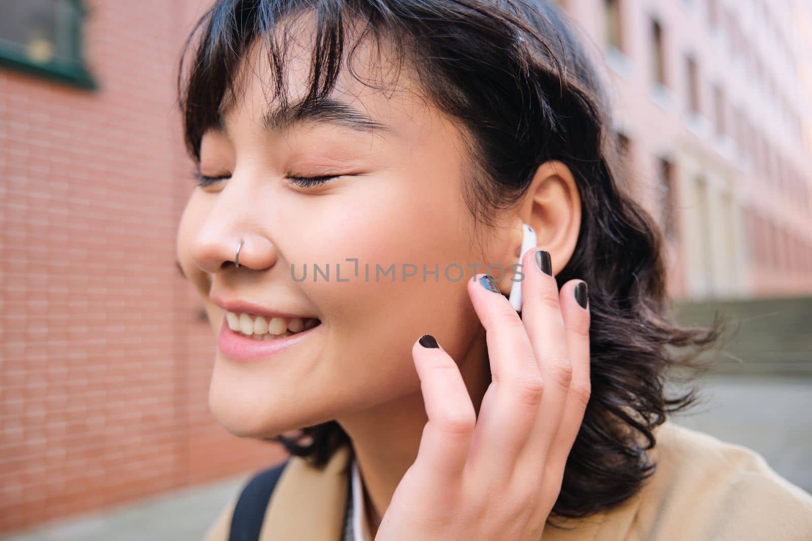 Close up portrait of young korean woman touches earphone, listens music in headphones with pleased smiling face, walks in city centre along the street.