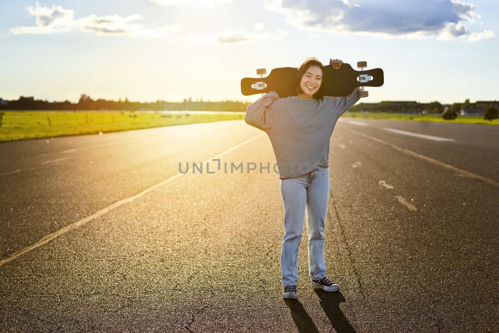 Young asian woman standing with longboard on sunny road, skating in skate park on her cruiser by Benzoix