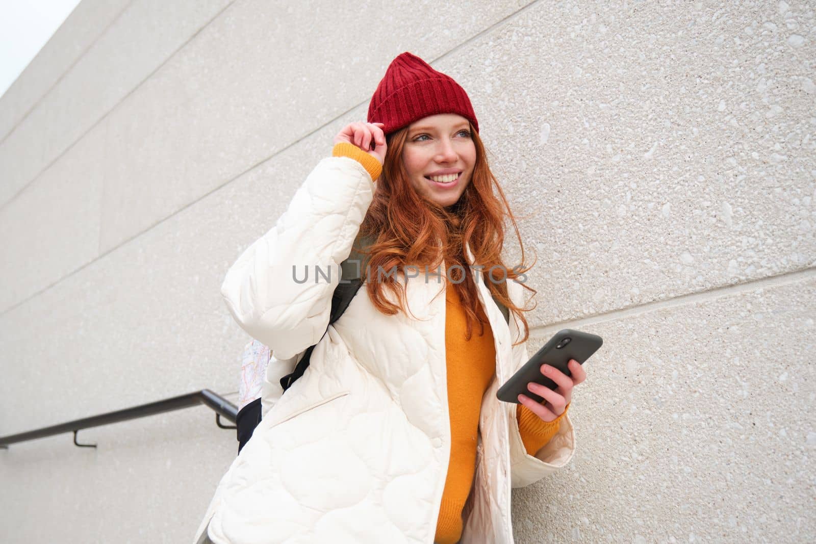 Stylish redhead woman in red hat, leans on wall on street, holds smartphone, uses internet phone app, reads message. Copy space