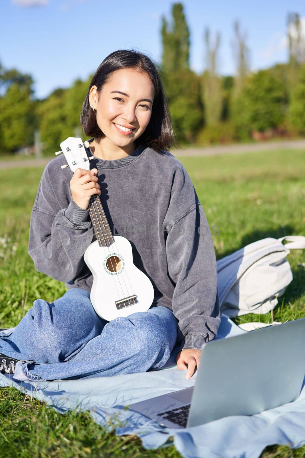 Smiling asian girl learns how to play ukulele on laptop, video chat with music teacher, sitting with instrument in park on grass.