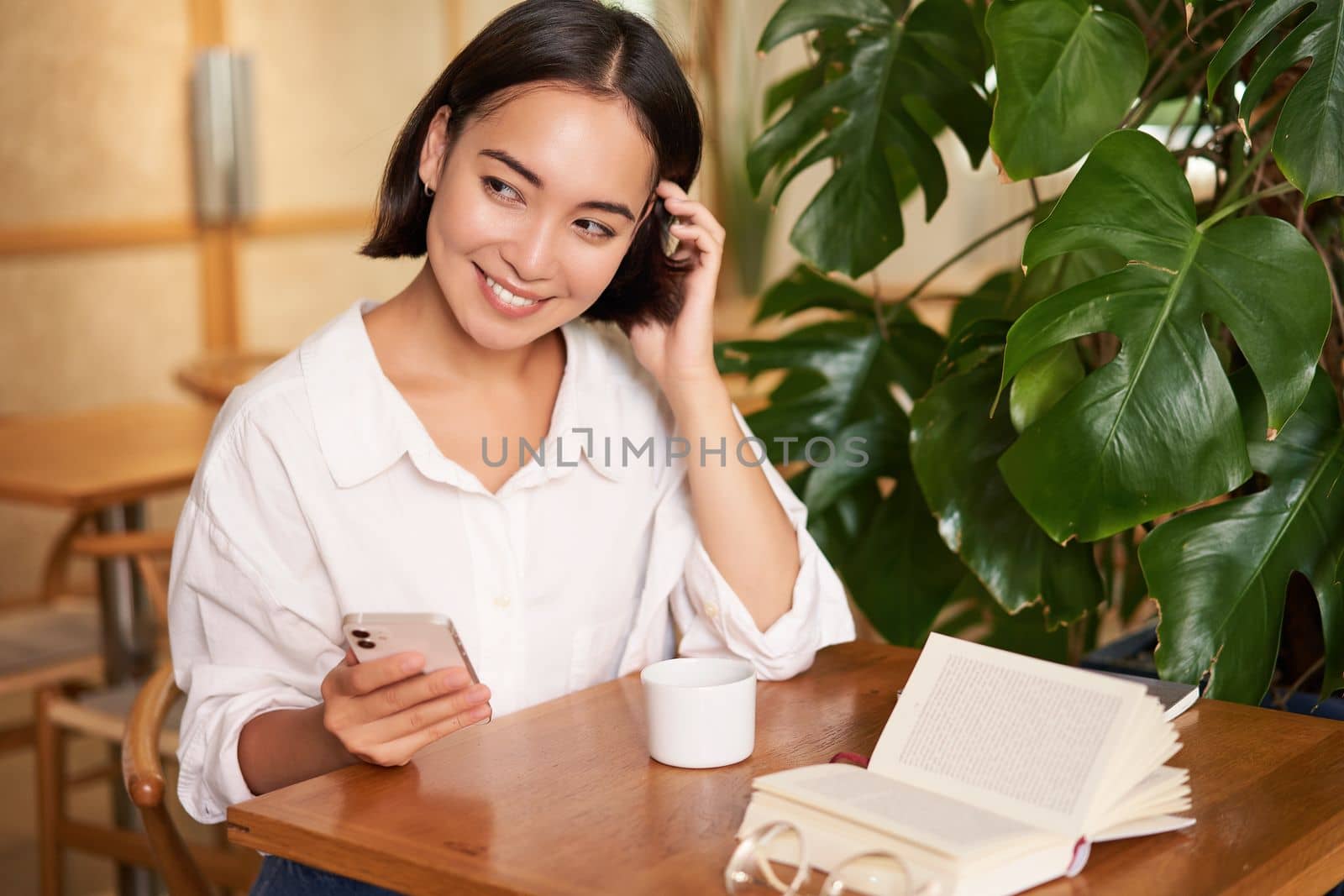 Beautiful young woman in cafe with cup of coffee and book, holding smartphone, using mobile phone, paying contactless waiting for waiter.