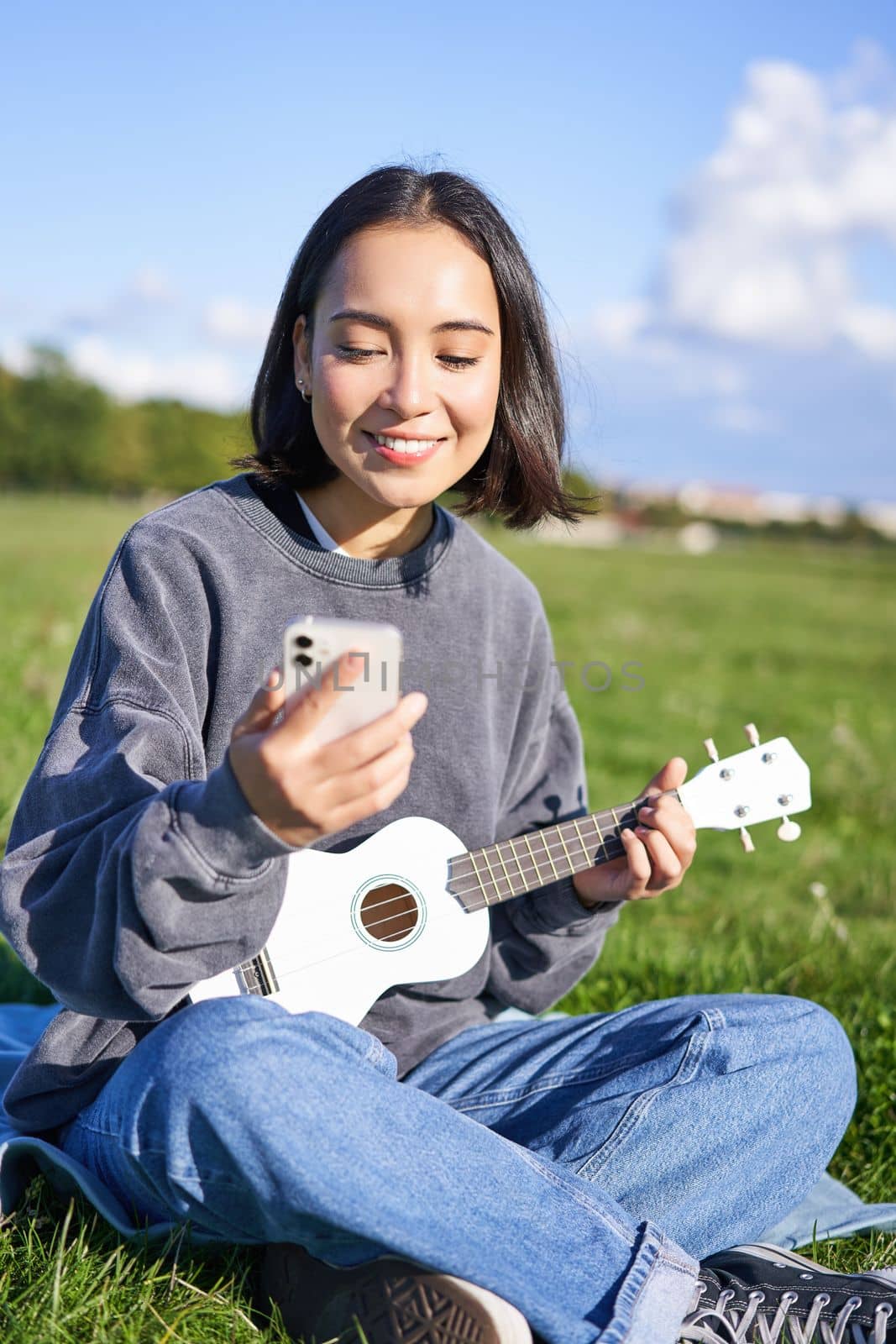 Smiling asian girl looking at guitar app, singing and playing ukulele while peeking at chords on smartphone, sitting outdoors in park on blanket.