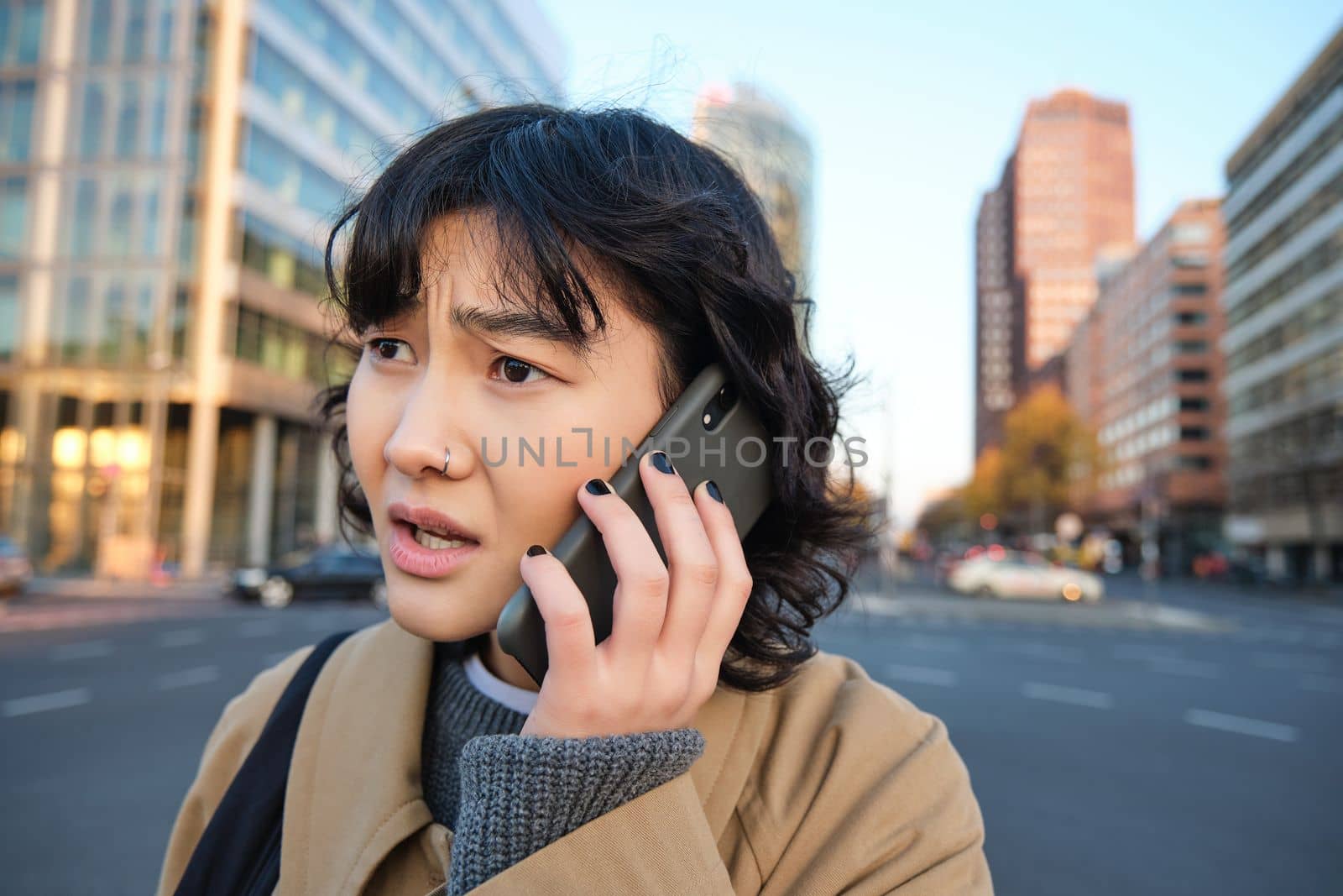 Close up portrait of concerned asian woman, talks on mobile phone and hears bad news, looks worried, feels frustrated by telephone conversation, stands on street.