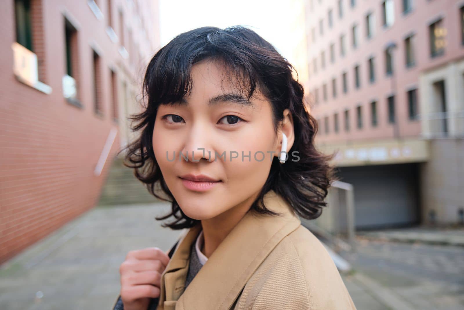 Stylish modern asian girl, student in wireless headphones, walks around city with backpack, smiling with happy face. Copy space