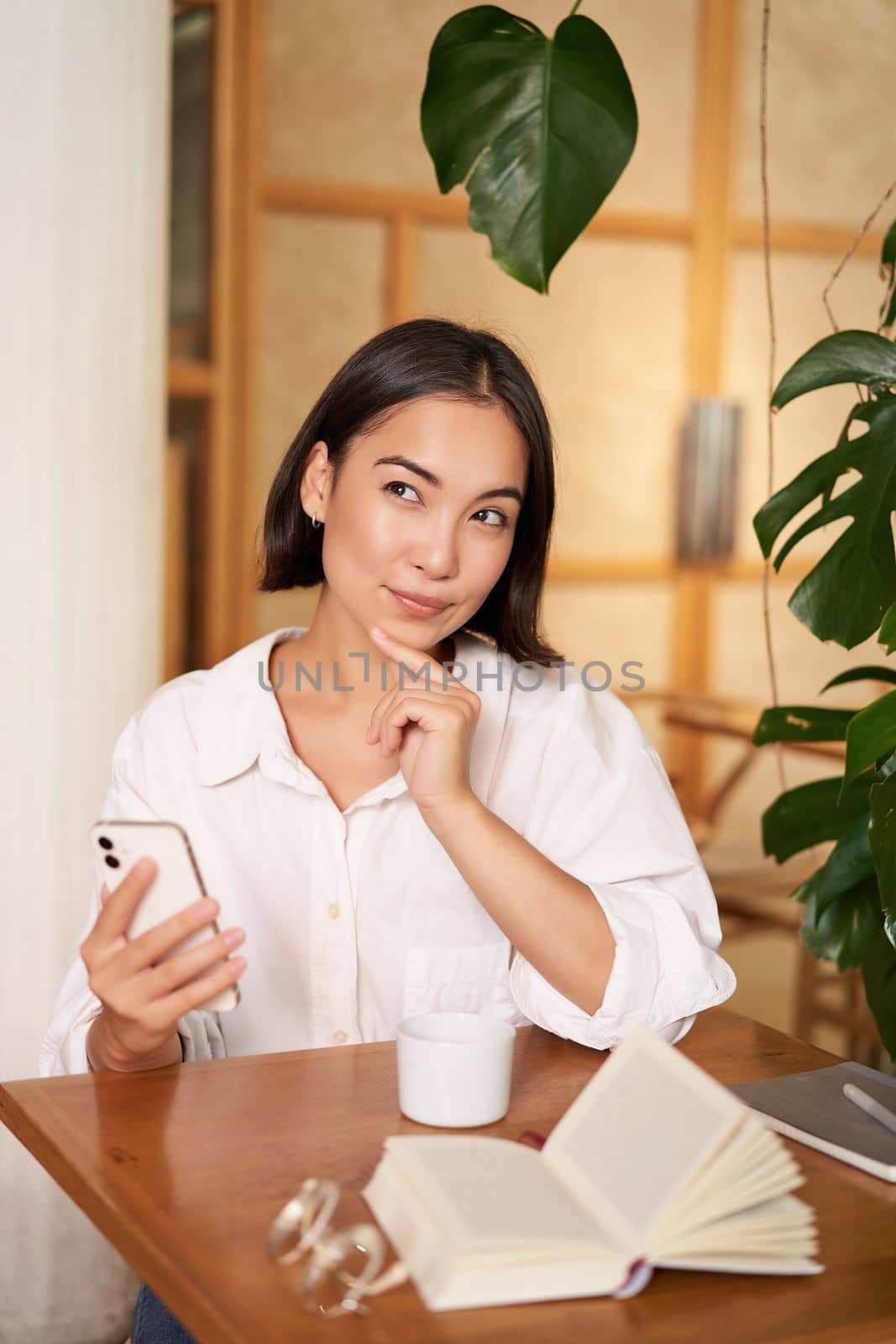 Beautiful young woman, 25 years old, thinking, holding smartphone and looking thoughtful, sitting in cafe, deciding smth.