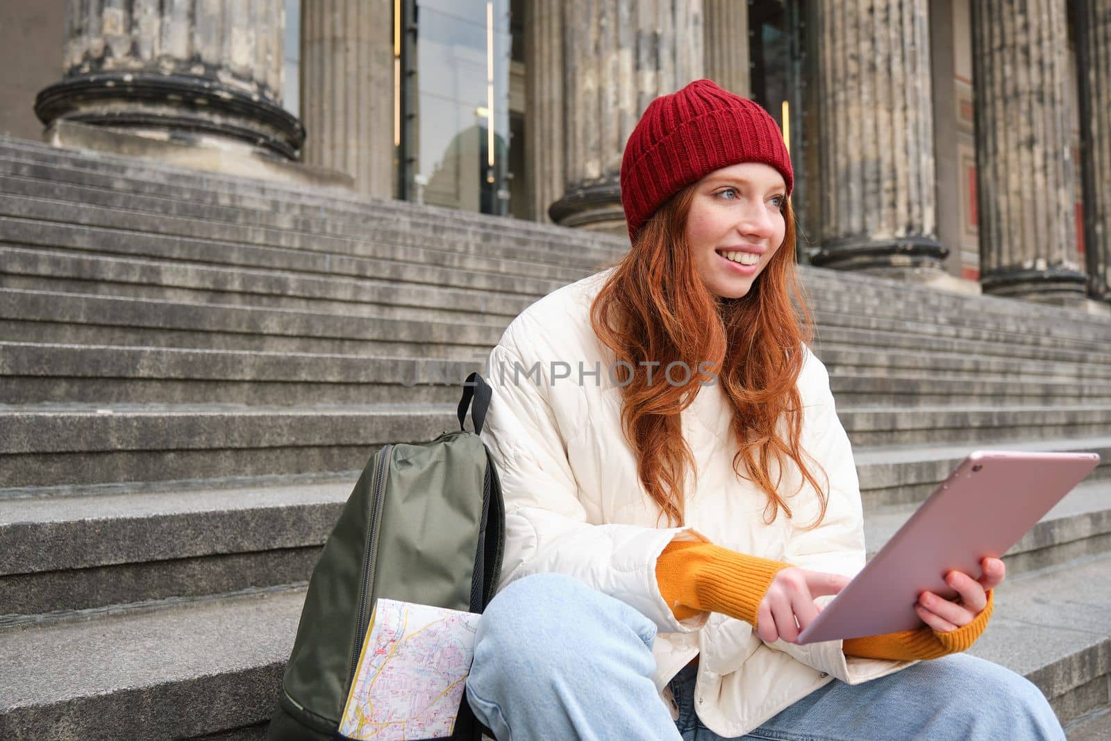 Beautiful young modern girl with red hair, holds digital tablet, sits on stairs near museum and connects public internet, sends message on gadget app.