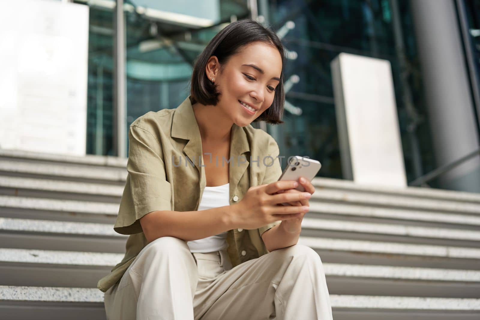 Technology and communication. Young smiling girl, asian woman sits with smartphone, reads message with big smile.