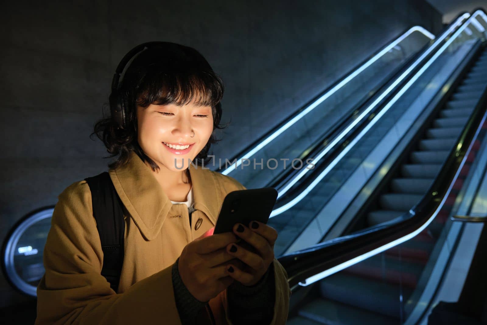 Portrait of young smiling asian girl, stands near escalator and waits for someone, reads message, checks map on smartphone, listens music in headphones.