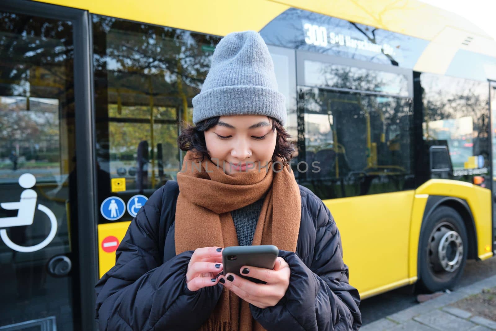 Image of girl student waiting for public transport, checks schedule on smartphone app, stands near city bus. Urban lifestyle concept