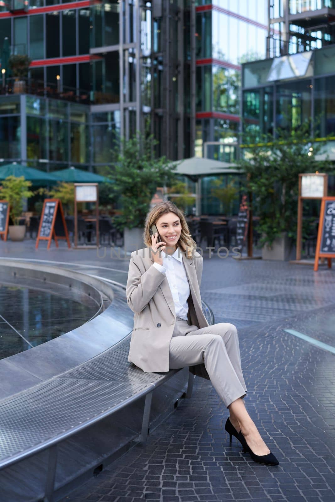 Vertical shot of young businesswoman in suit, sitting casually outside office buldings, talking on smartphone, making a phone call and smiling.