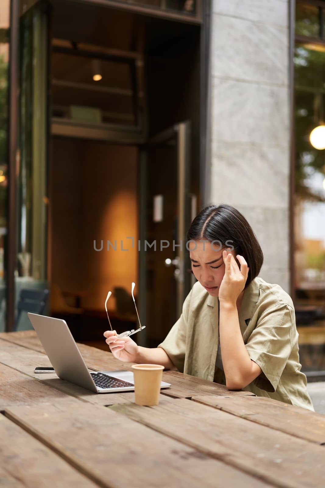 Young asian woman feeling tired after working with laptop, sitting in cafe on bench outdoors, drinking coffee, looking exhausted.