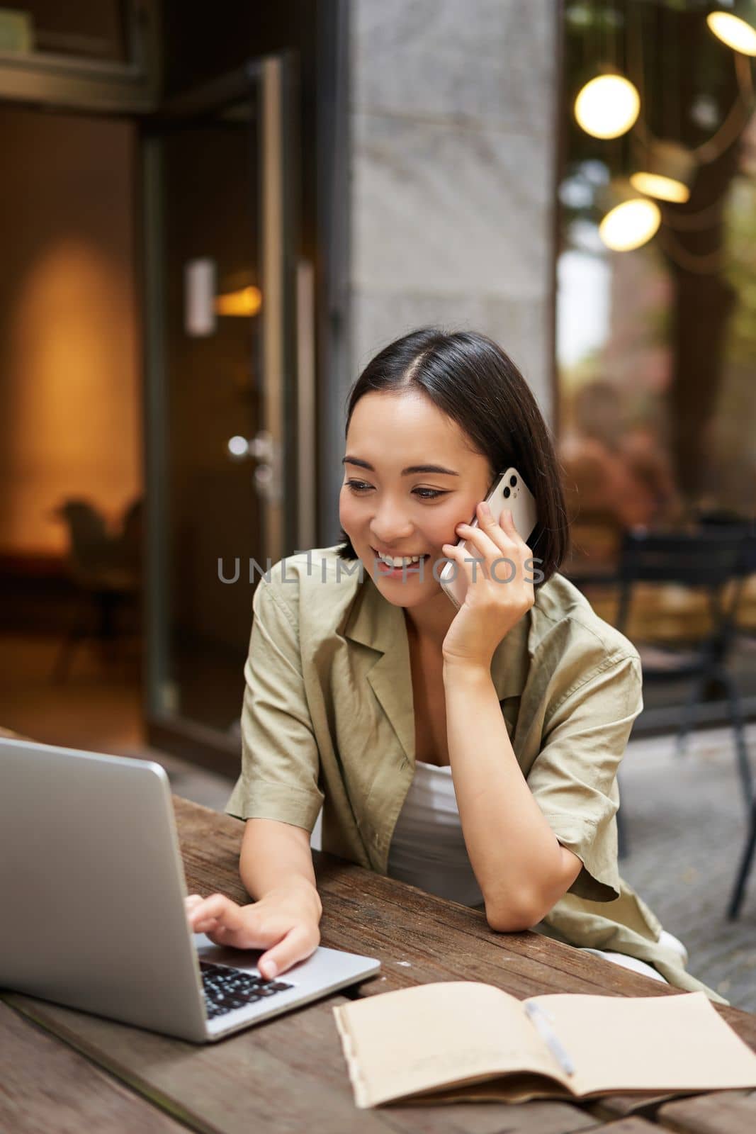 Happy girl calling on mobile phone, talking on smartphone and working with laptop, doing homework, studying outdoors from cafe.